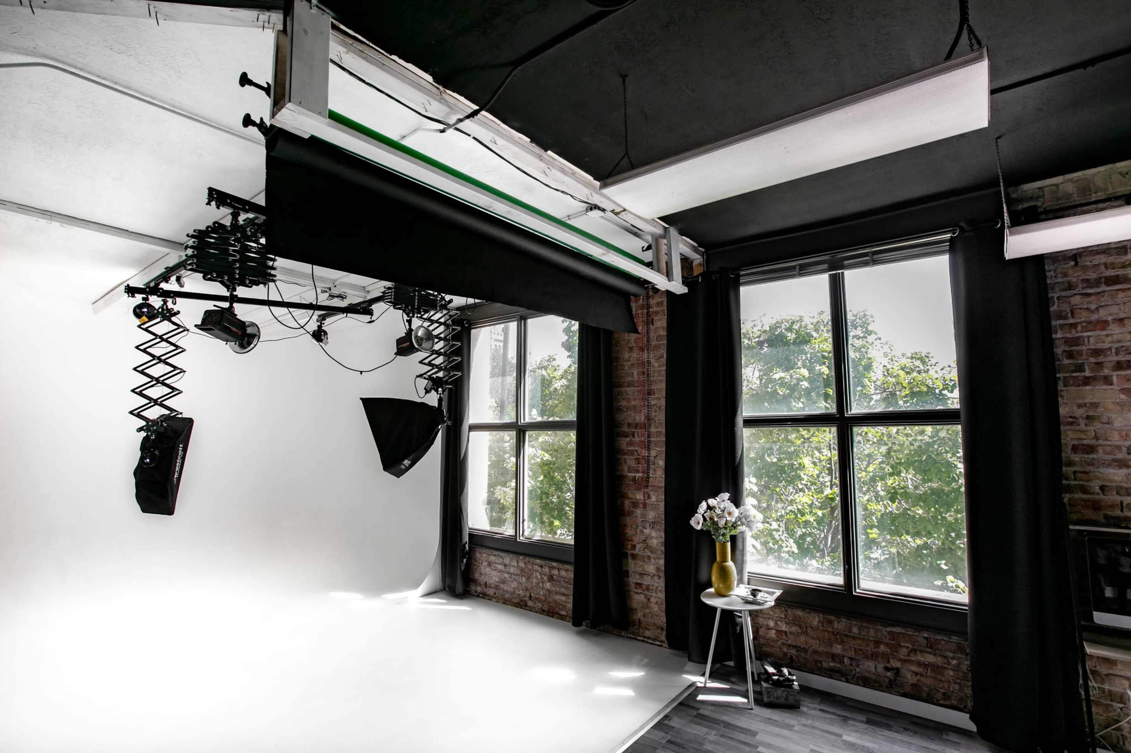 A photography studio with a white backdrop, overhead lighting equipment, and large windows surrounded by black curtains.