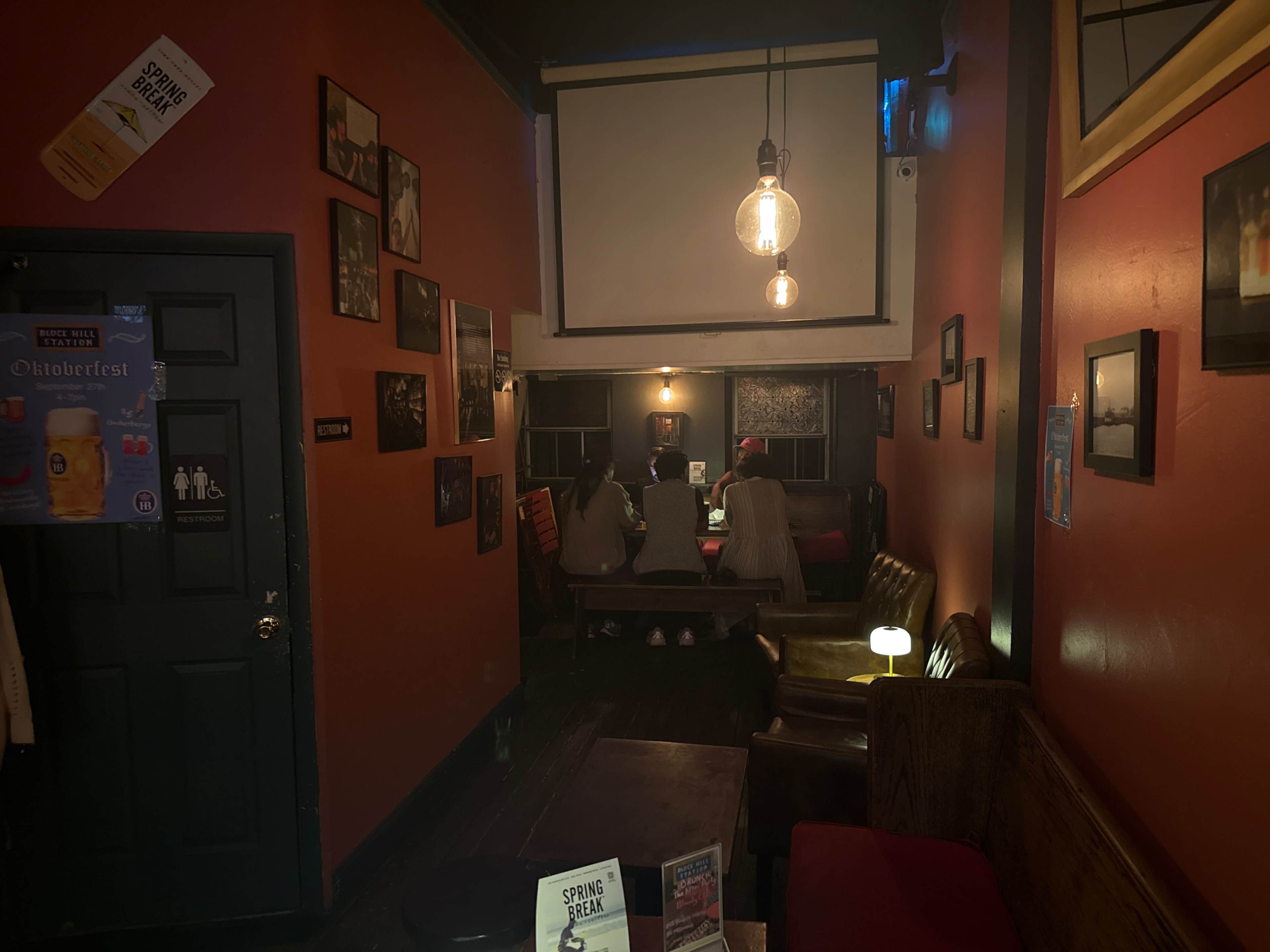The image shows a dimly lit café with orange walls, featuring a few tables and framed photographs, where two people are seated at a table in the background.