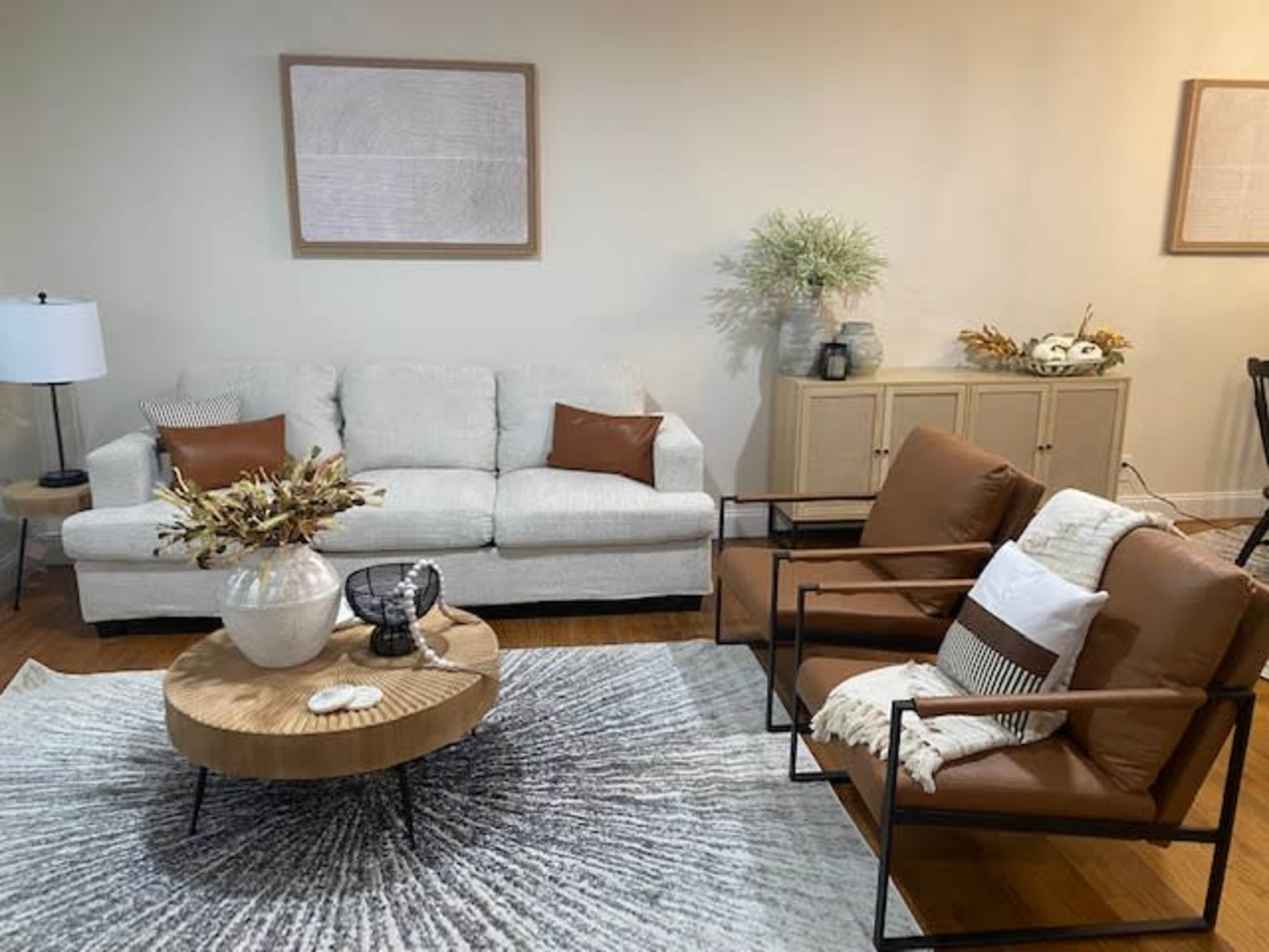 A living room with a light-colored sofa, two brown chairs, a round coffee table, and decorative elements on a large circular rug.