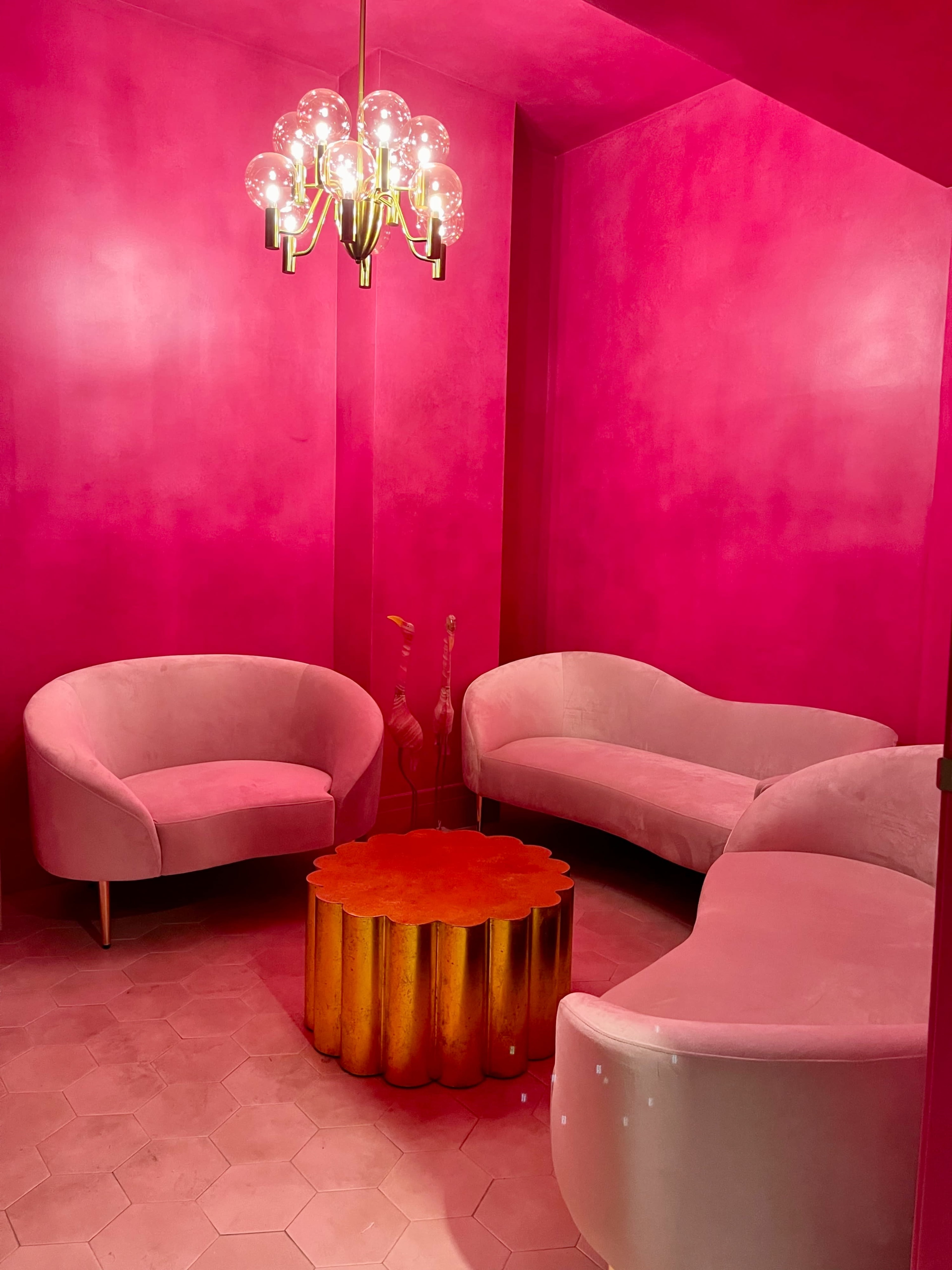 A room with bright pink walls, featuring two curved sofas and a round gold table in the center, illuminated by a modern chandelier.
