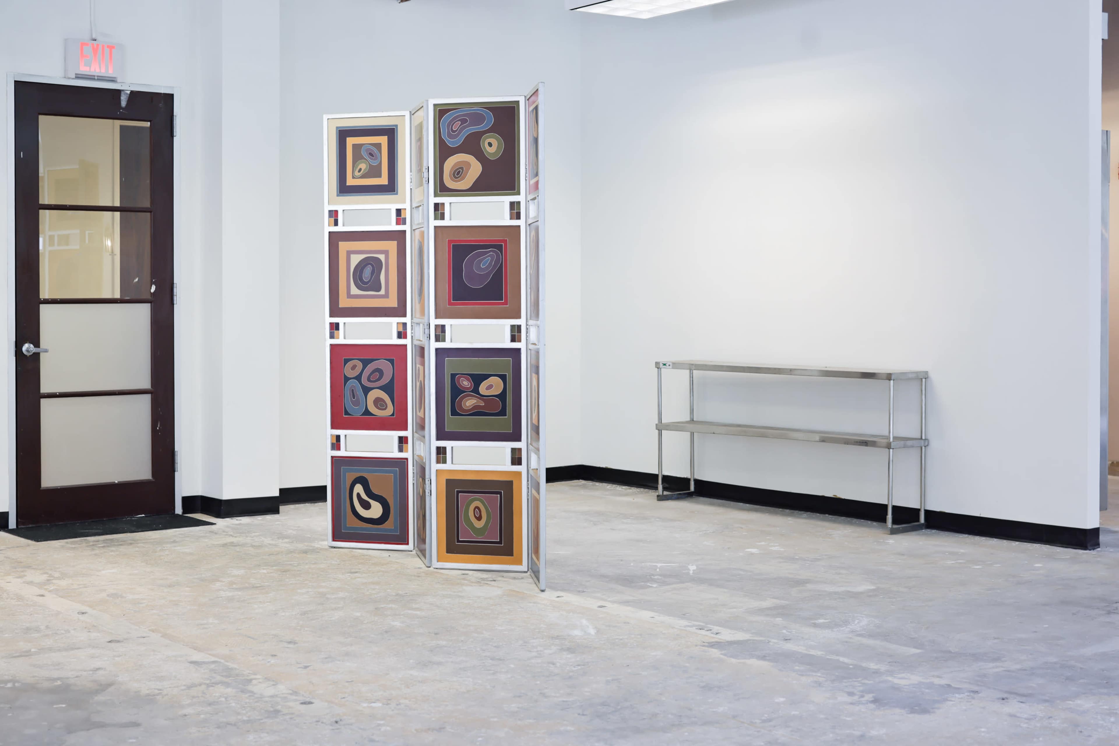 The image shows a sparsely furnished art gallery with a partition displaying colorful abstract artworks and an empty metal shelf against a plain wall.