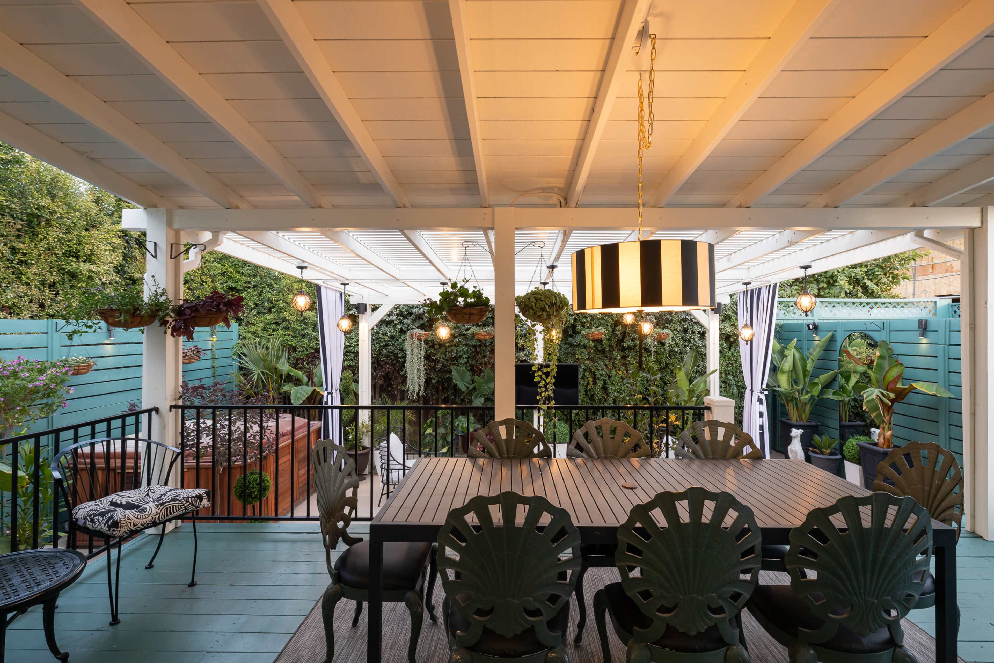 A spacious outdoor dining area features a large table surrounded by patterned chairs, with hanging lights and a view of a garden.