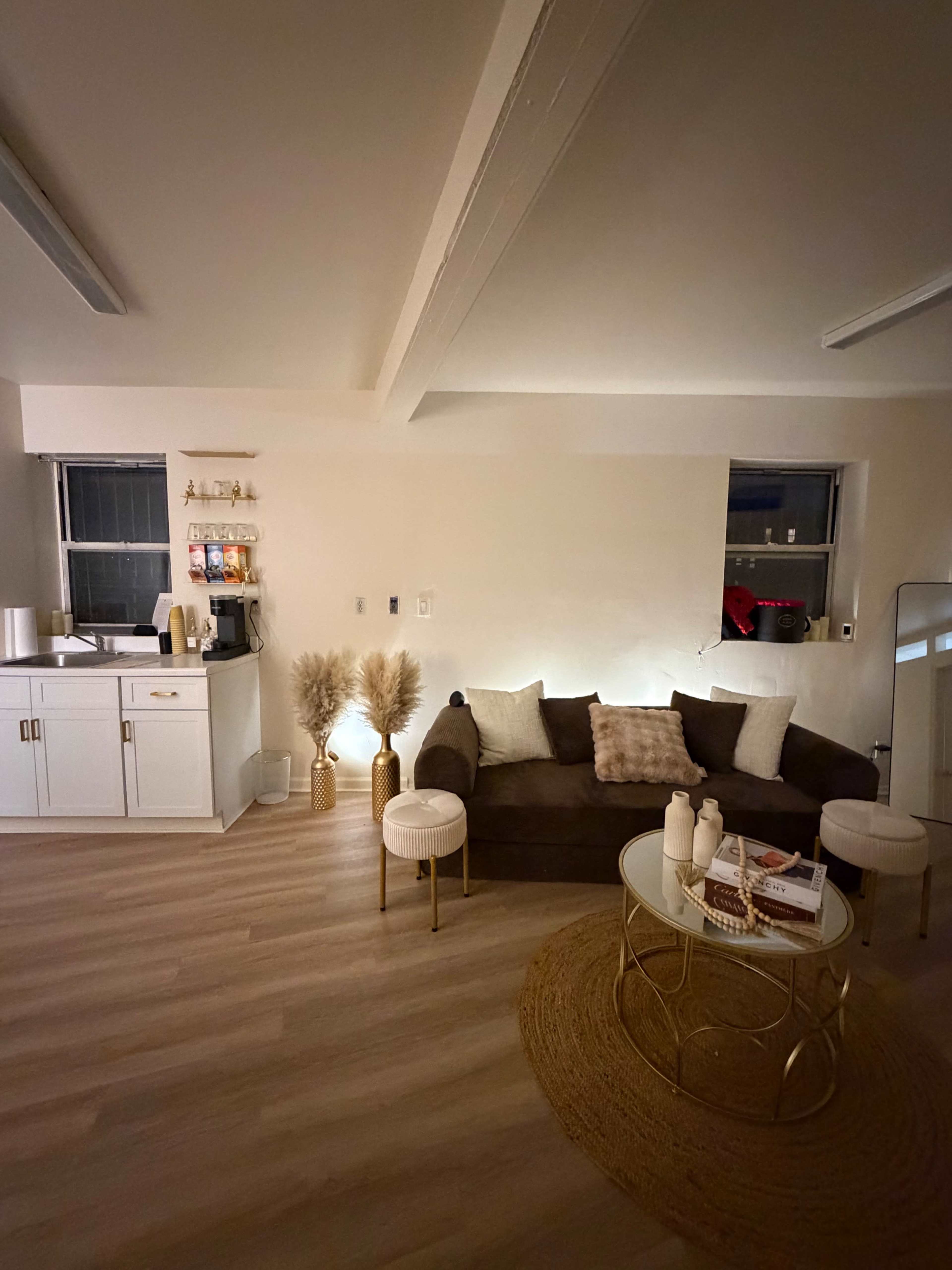 The image shows a cozy, softly lit living area featuring a dark sofa, two white side tables, a round rug, and a kitchenette in the background.