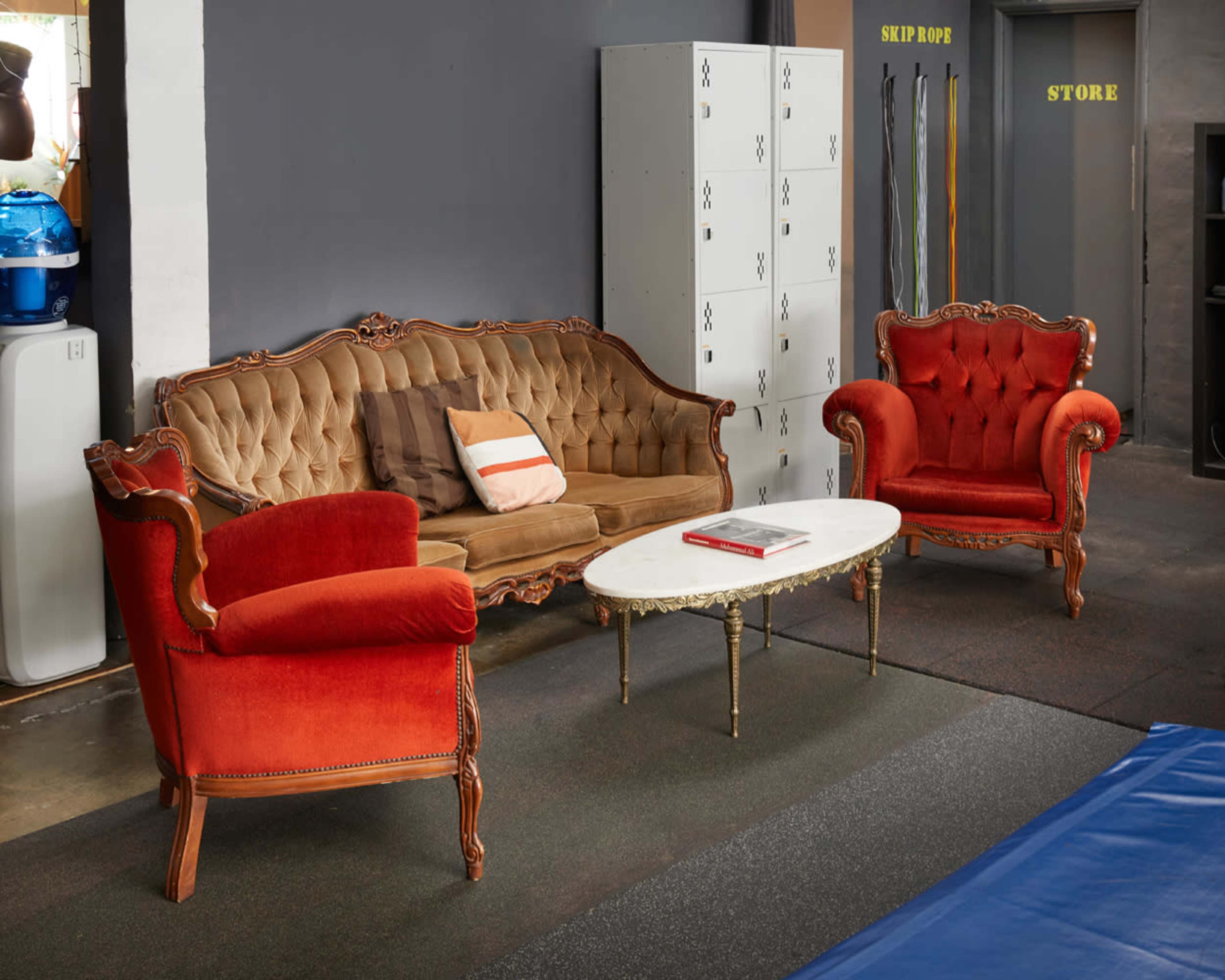 A vintage seating area with an ornate brown sofa and two red armchairs arranged around a white coffee table in a gym setting.