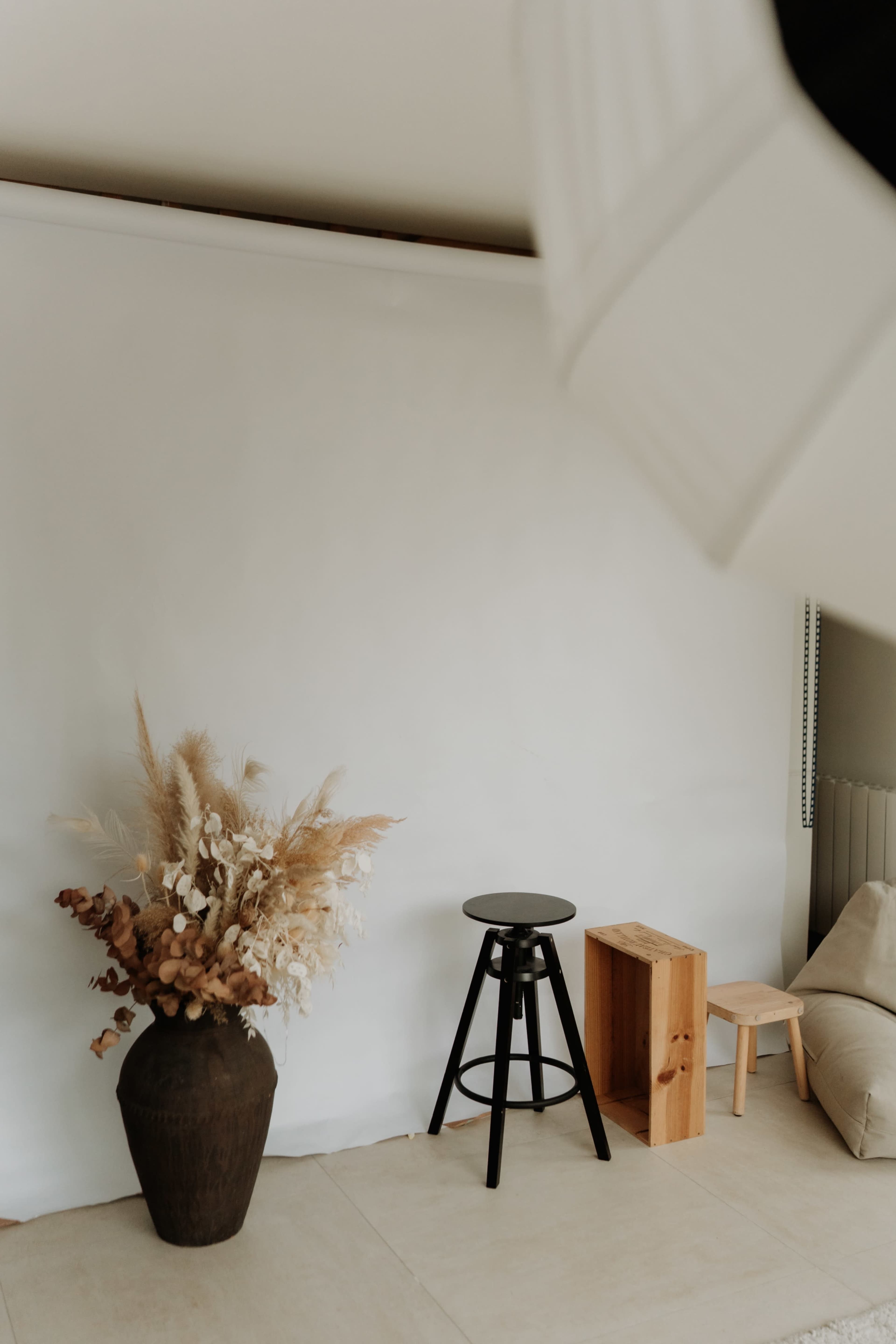The image shows a minimalistic interior setup featuring a black stool, a wooden box, and a vase of dried flowers against a white backdrop.