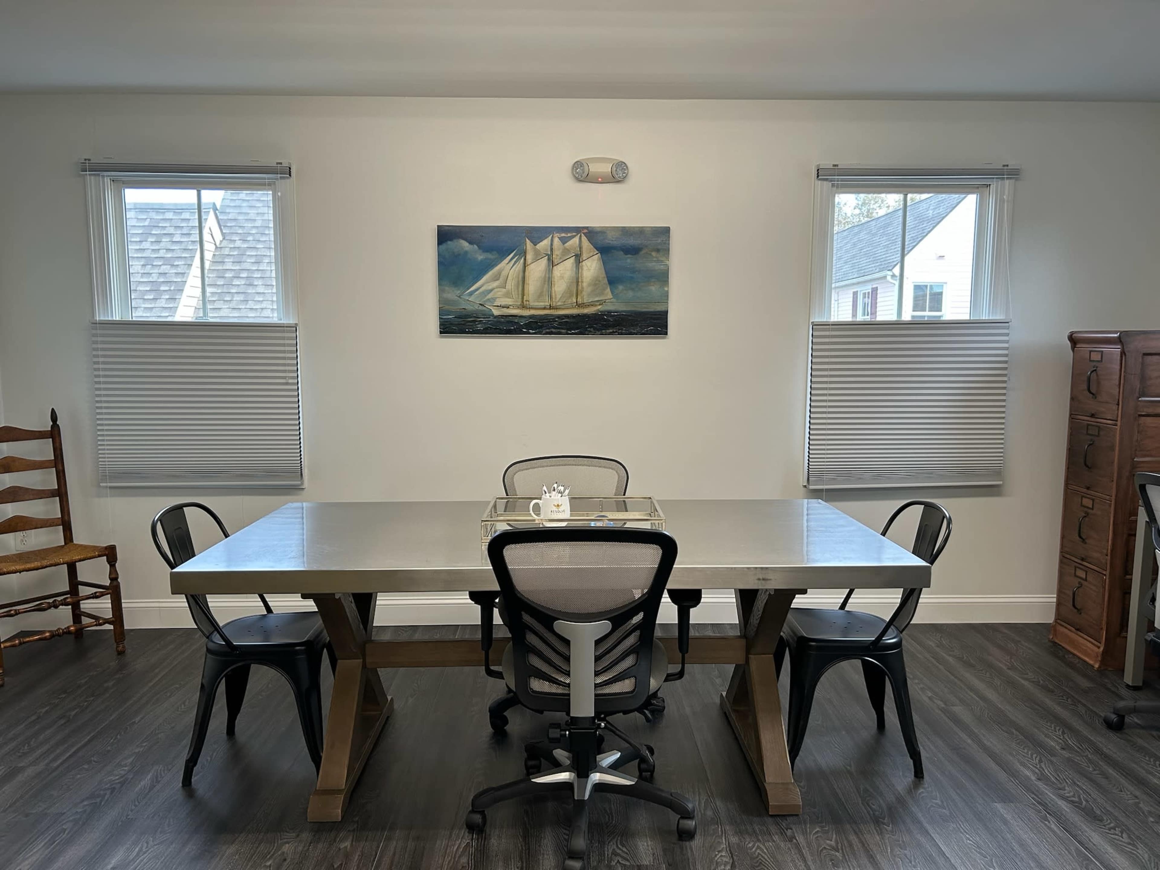 A spacious dining area features a large table with five chairs, framed by two windows with blinds and a painting of a sailing ship on the wall.
