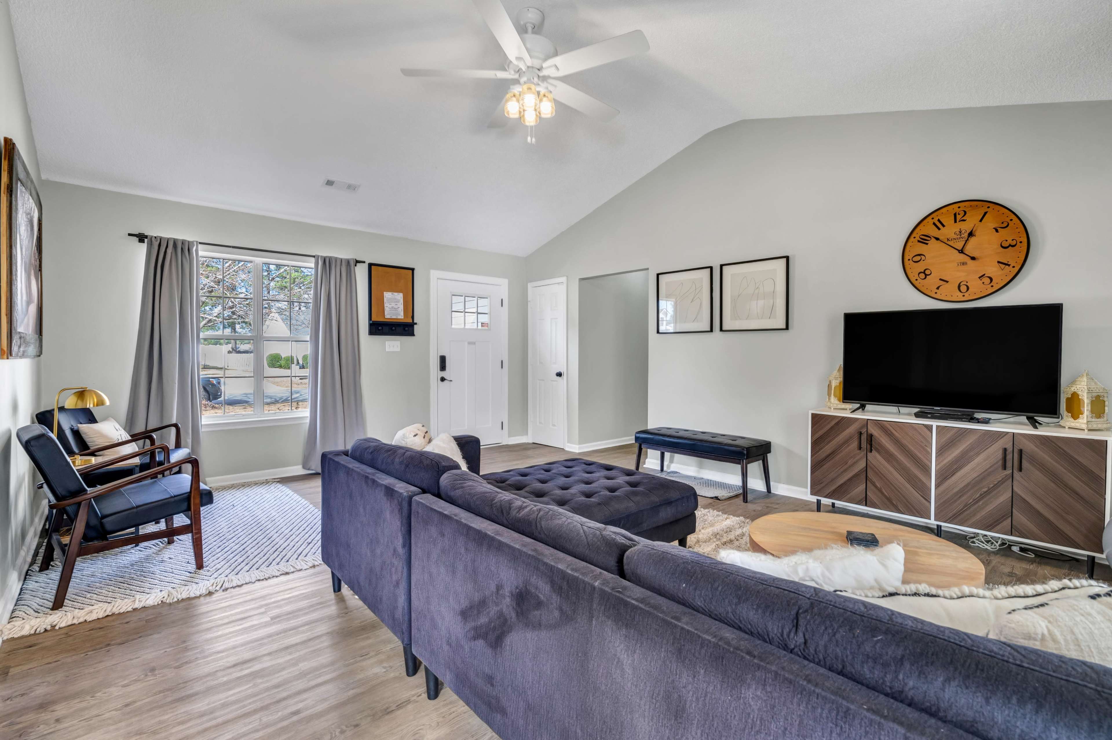 A modern living room features a sectional sofa, a coffee table, and a television on a low cabinet, illuminated by natural light from a large window.
