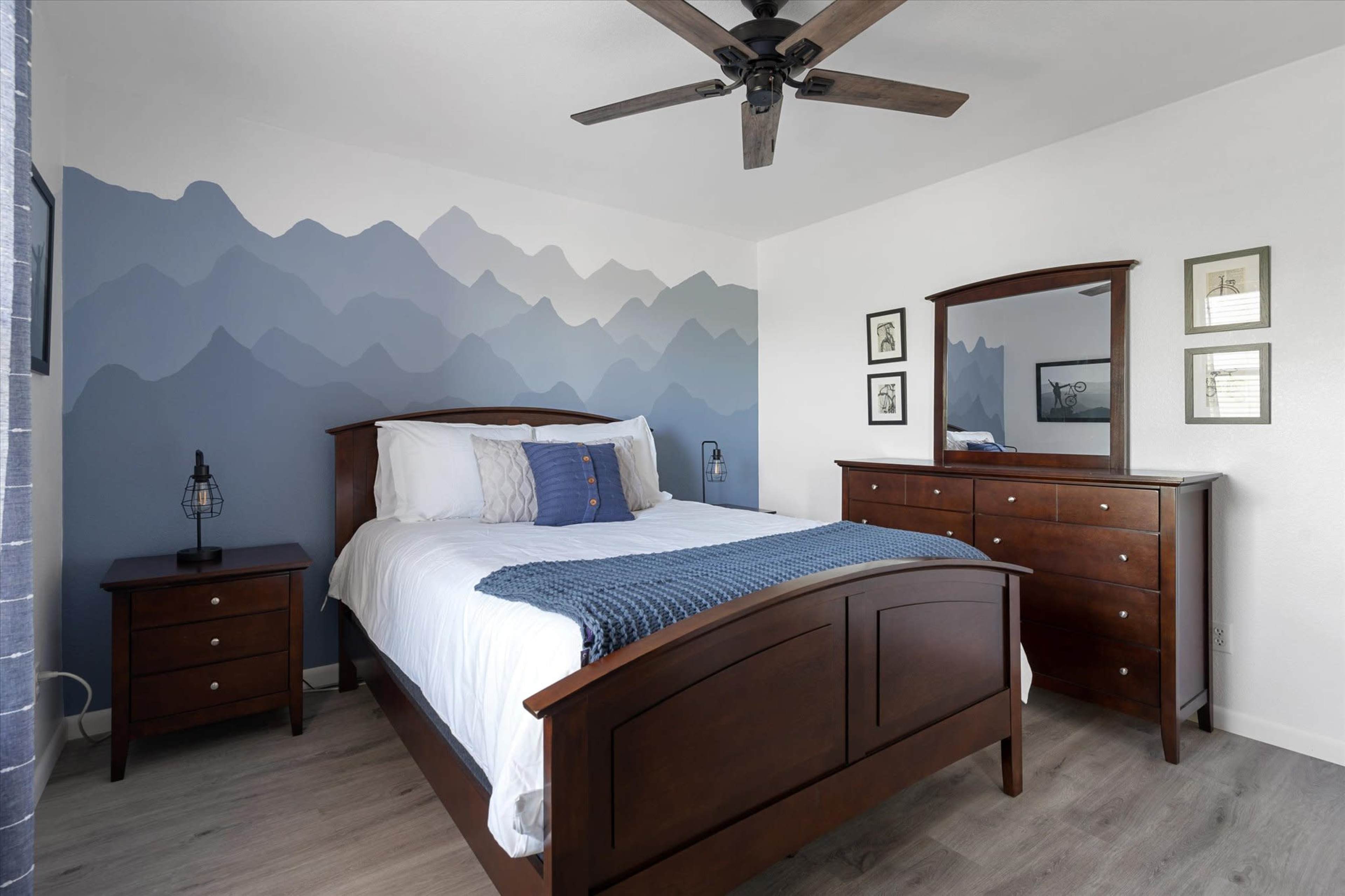 The image depicts a bedroom featuring a wooden bed frame, nightstand, and dresser, with a blue mountain mural on the wall behind the bed.