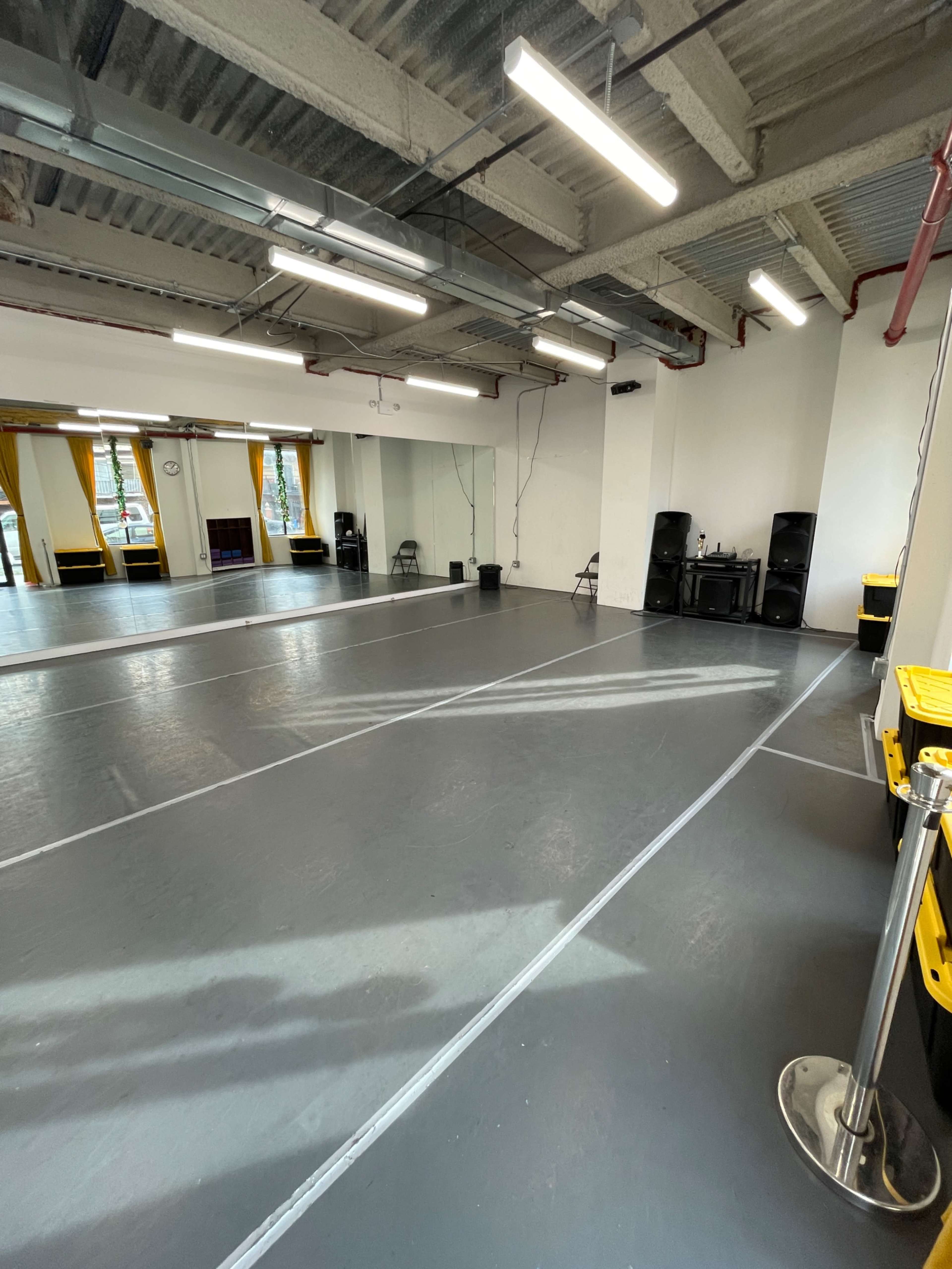 An empty dance studio features a smooth gray floor, mirrored walls, and fluorescent lighting.