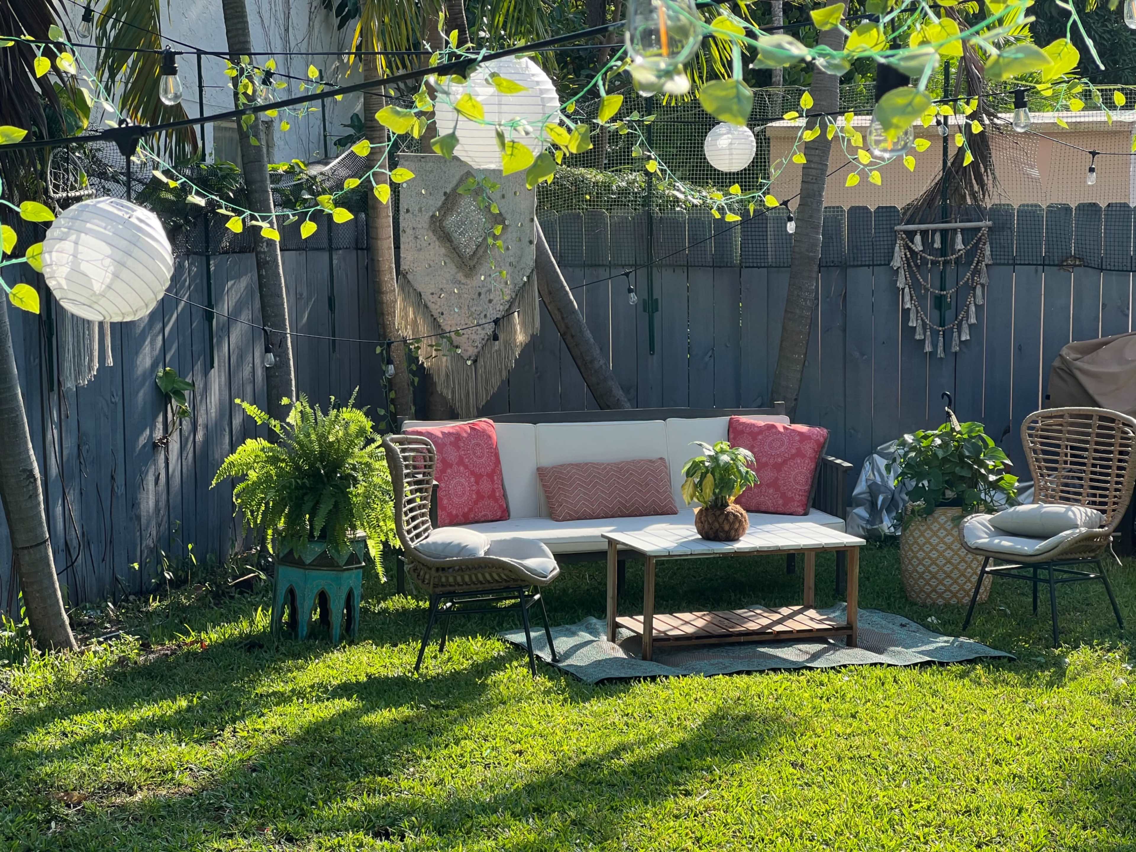 A cozy backyard seating area features a white sectional sofa, decorative pillows, a coffee table, and soft lighting among lush greenery.