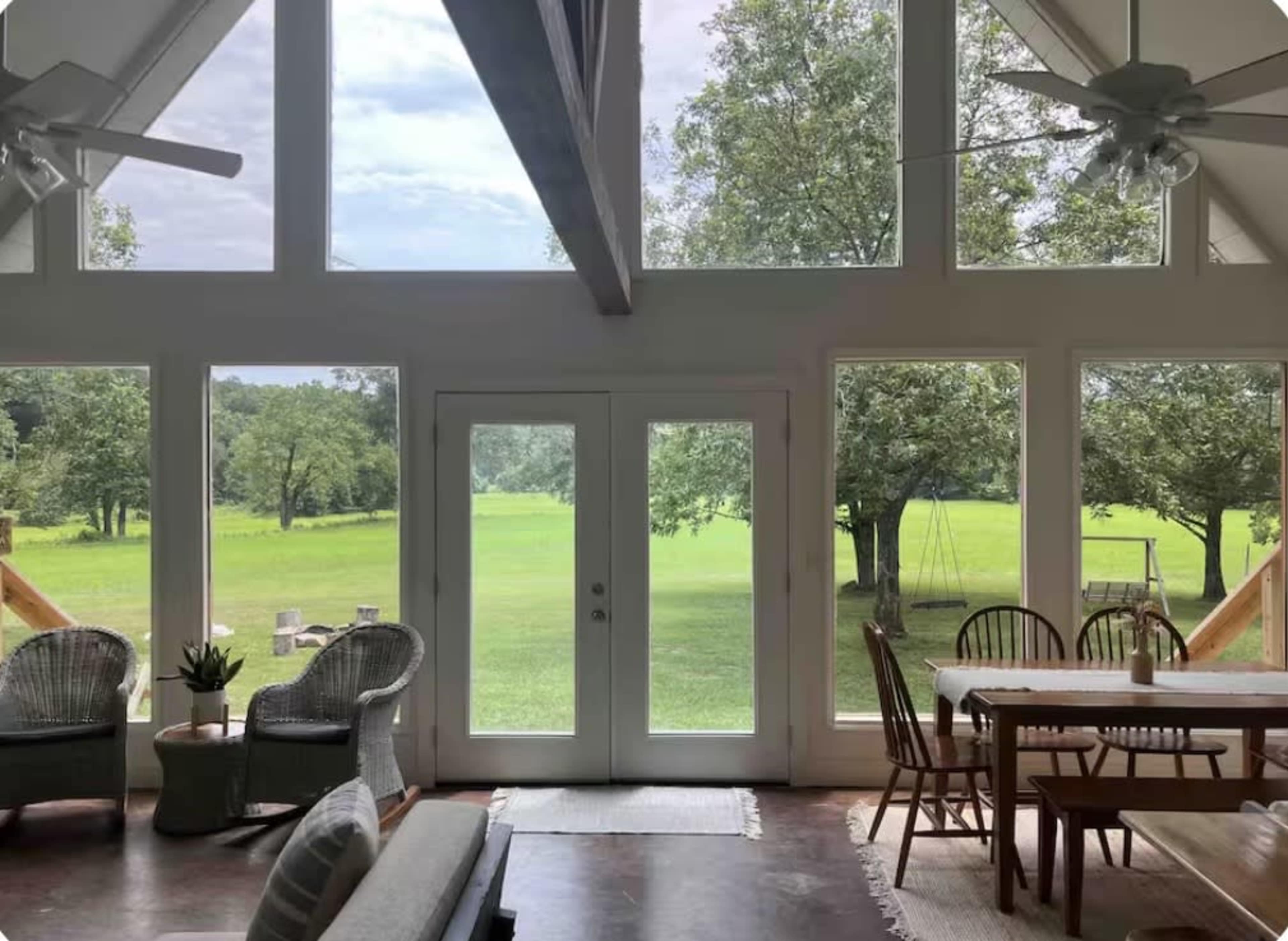 The image shows the interior of a modern home with large windows overlooking a green lawn and trees.