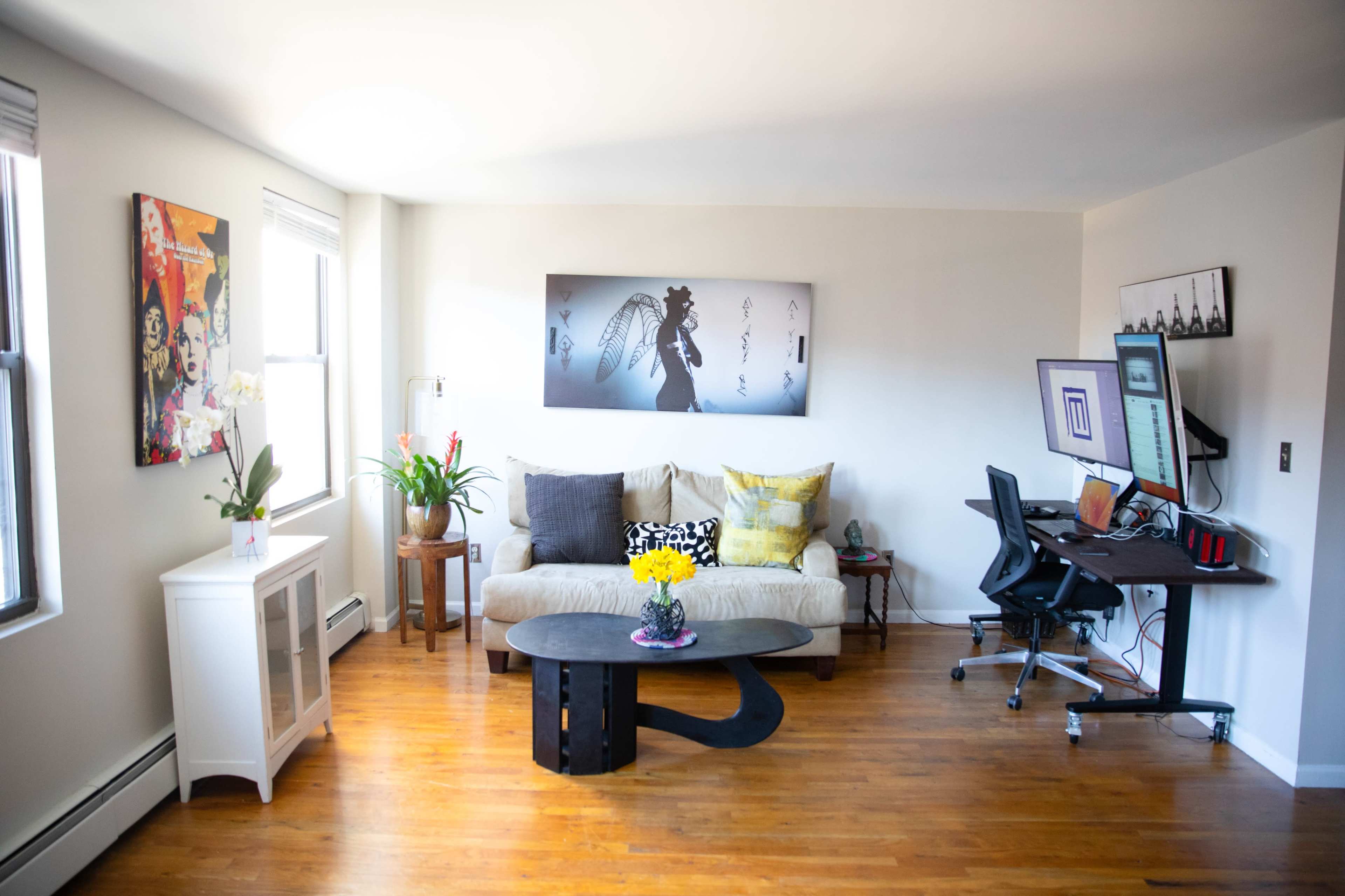 A bright living room features a beige couch with colorful pillows, a dark coffee table, a potted plant, and a modern desk setup with a computer and wall art.