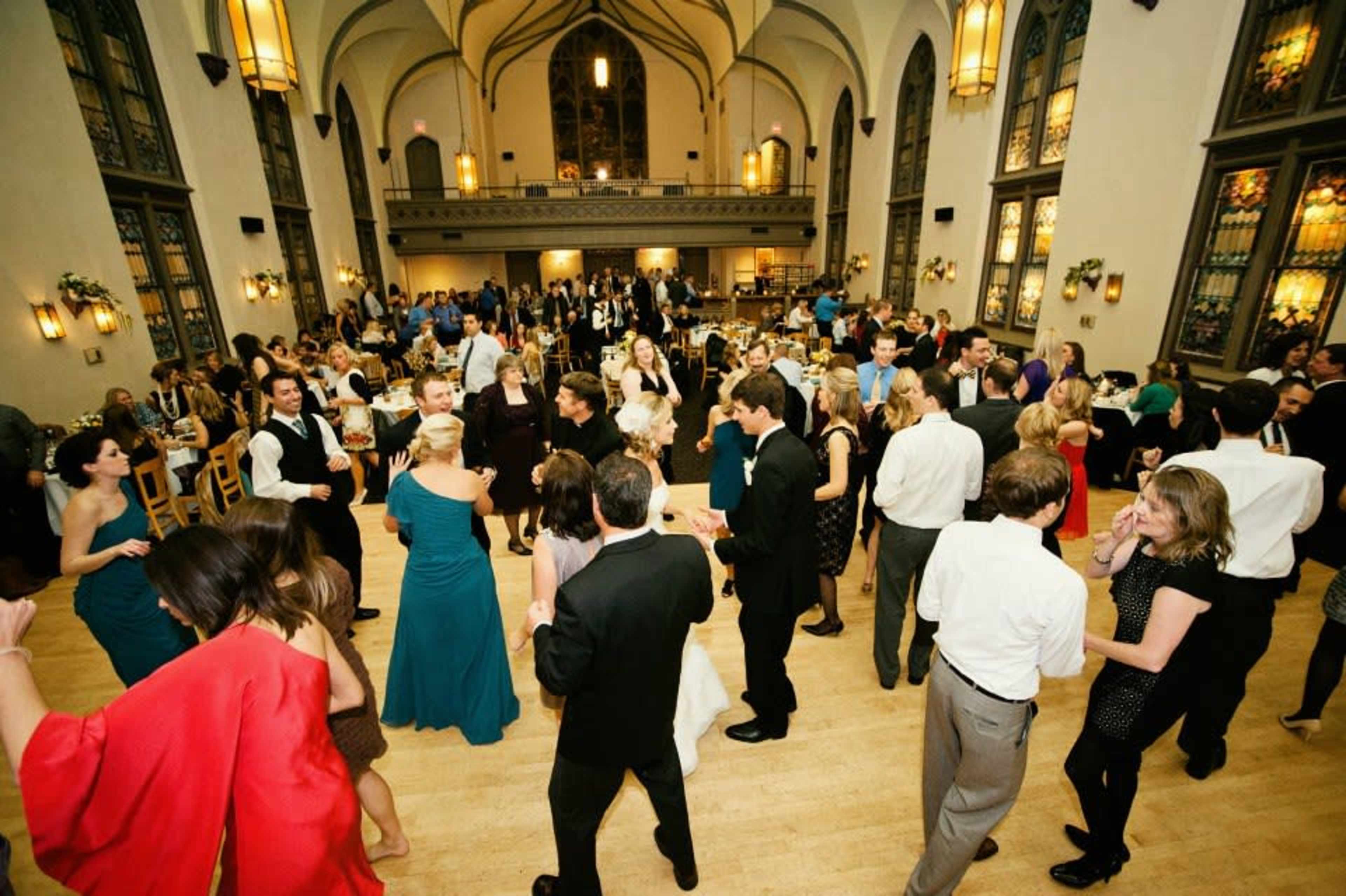 A large group of people is dancing and socializing in a spacious banquet hall with stained glass windows and elegant decor.
