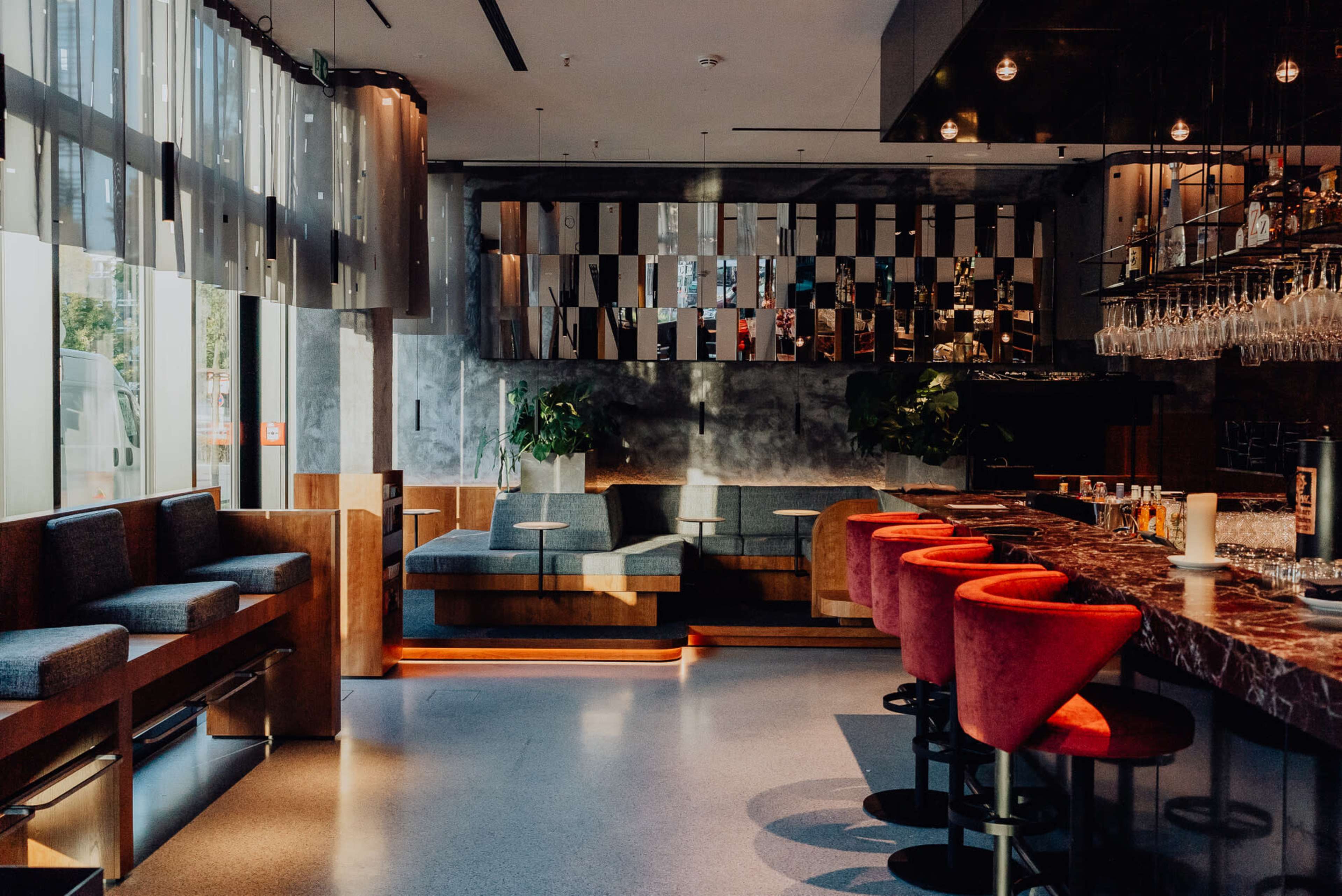 The image shows a modern bar with a wooden counter, colorful stools, and large windows allowing natural light to enter the space.