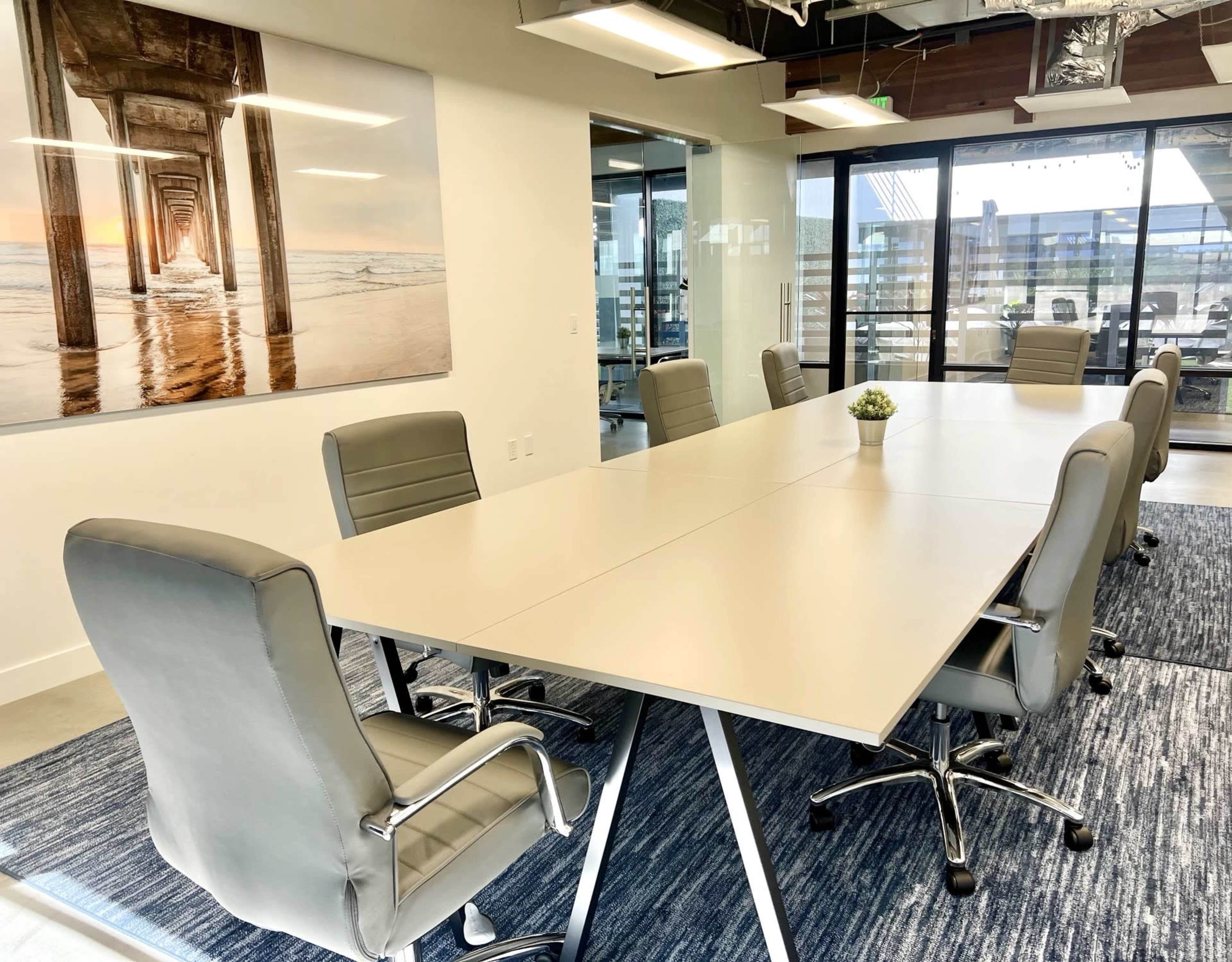 A modern conference room features a large, rectangular table surrounded by ergonomic chairs, with a seaside photograph on the wall and large windows overlooking an outdoor area.
