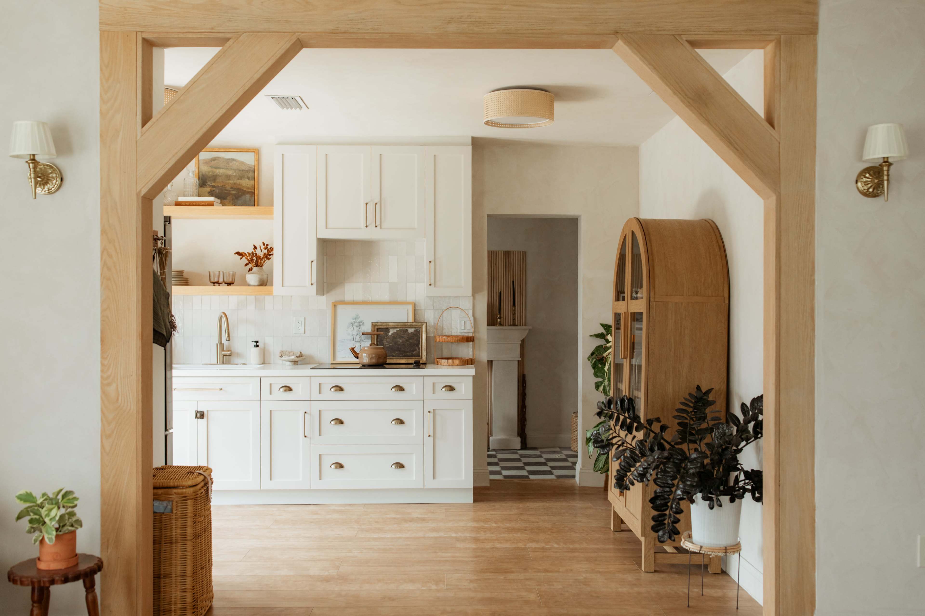 A bright kitchen with white cabinetry and a wooden archway opens up to a cozy dining area featuring plants and decorative furniture.