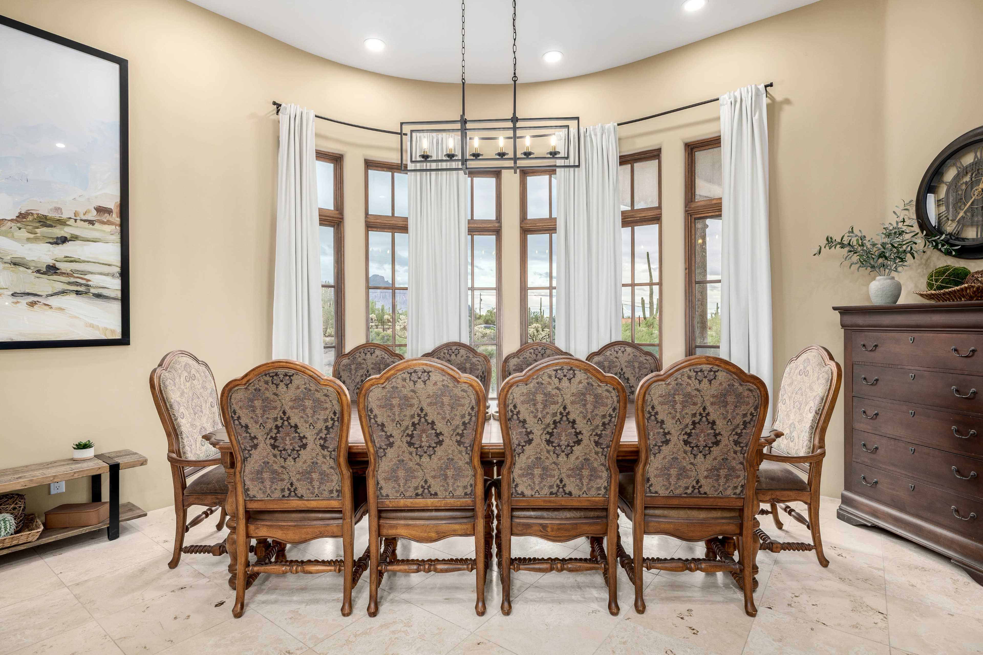 A large dining table with ornate chairs is set in a sunlit room featuring large windows and neutral-colored walls.