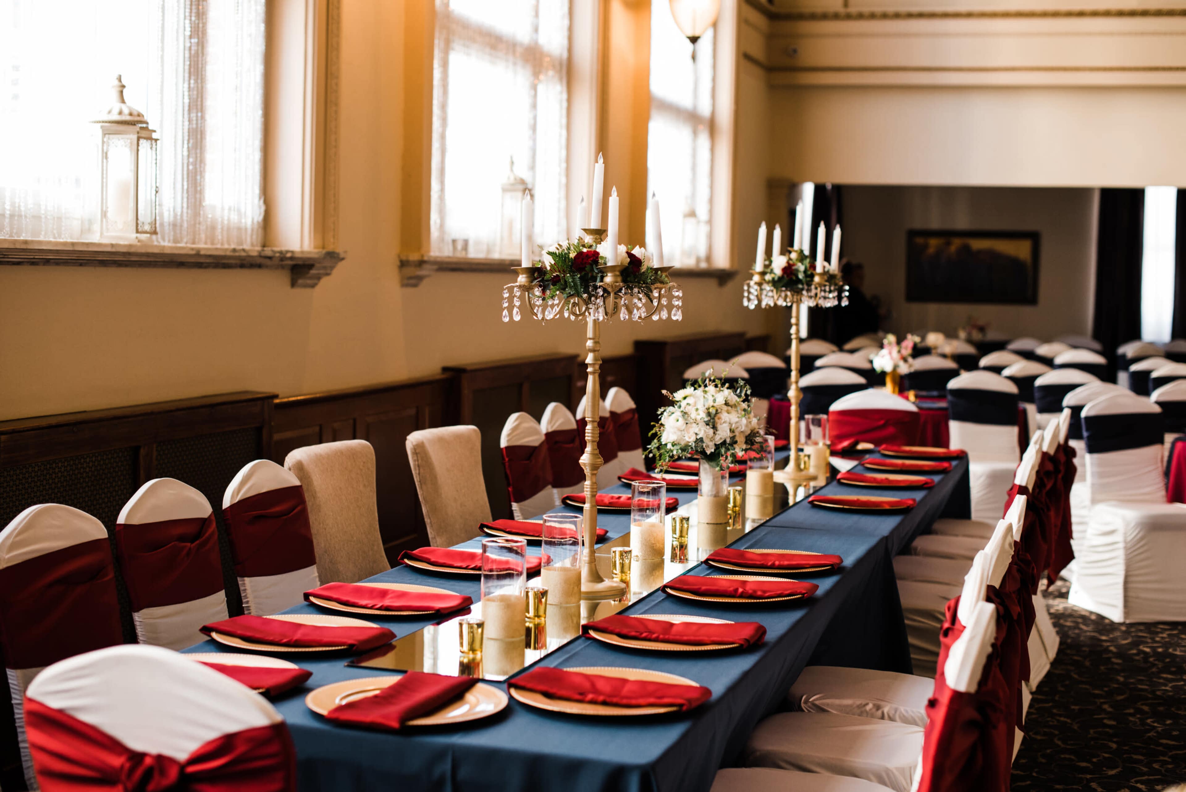 A long dining table is set with blue and red tableware, surrounded by decorated chairs in a banquet hall with large windows.