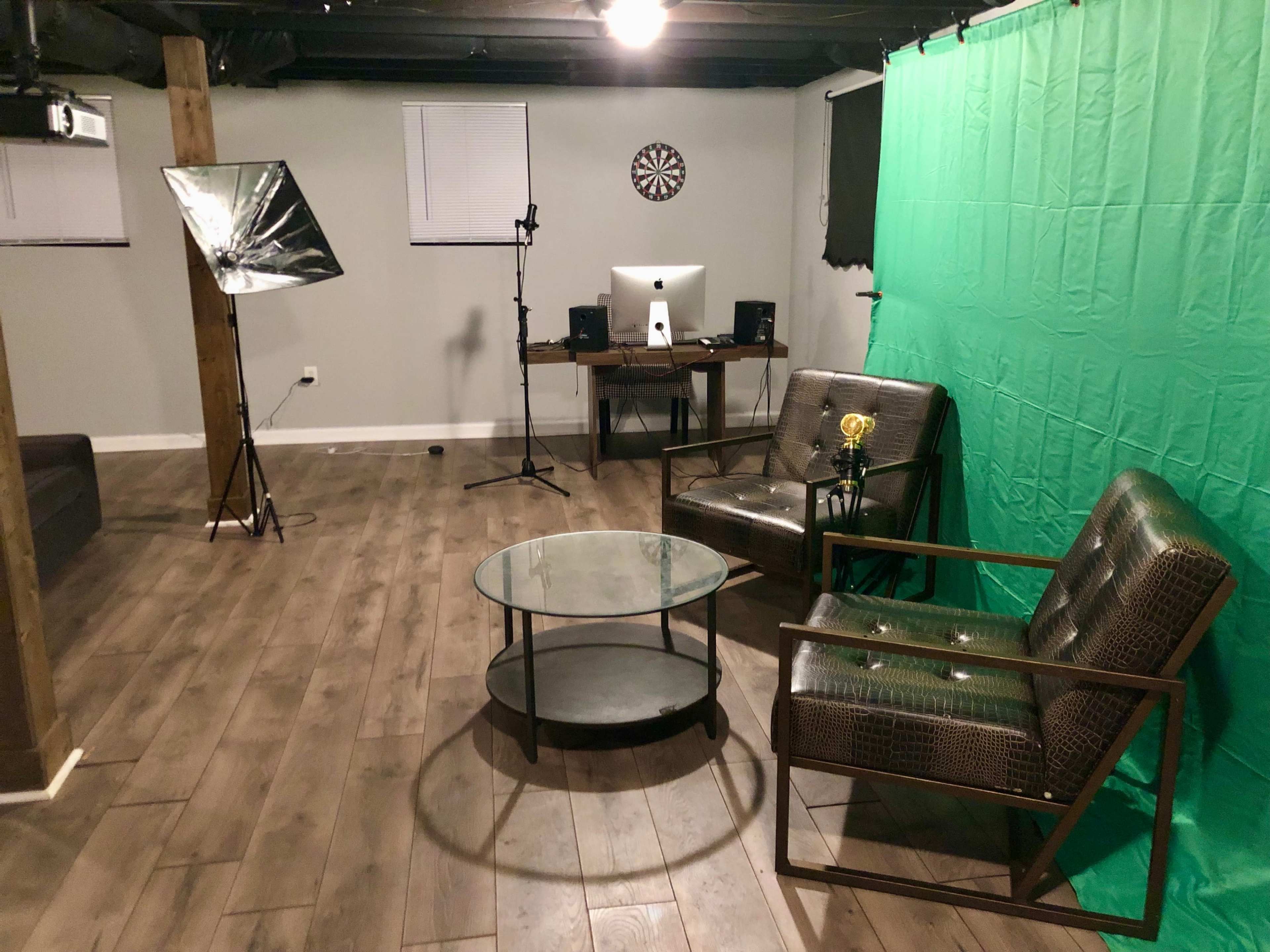 A basement studio setup with a green screen, two chairs, a glass coffee table, and equipment for recording or broadcasting.