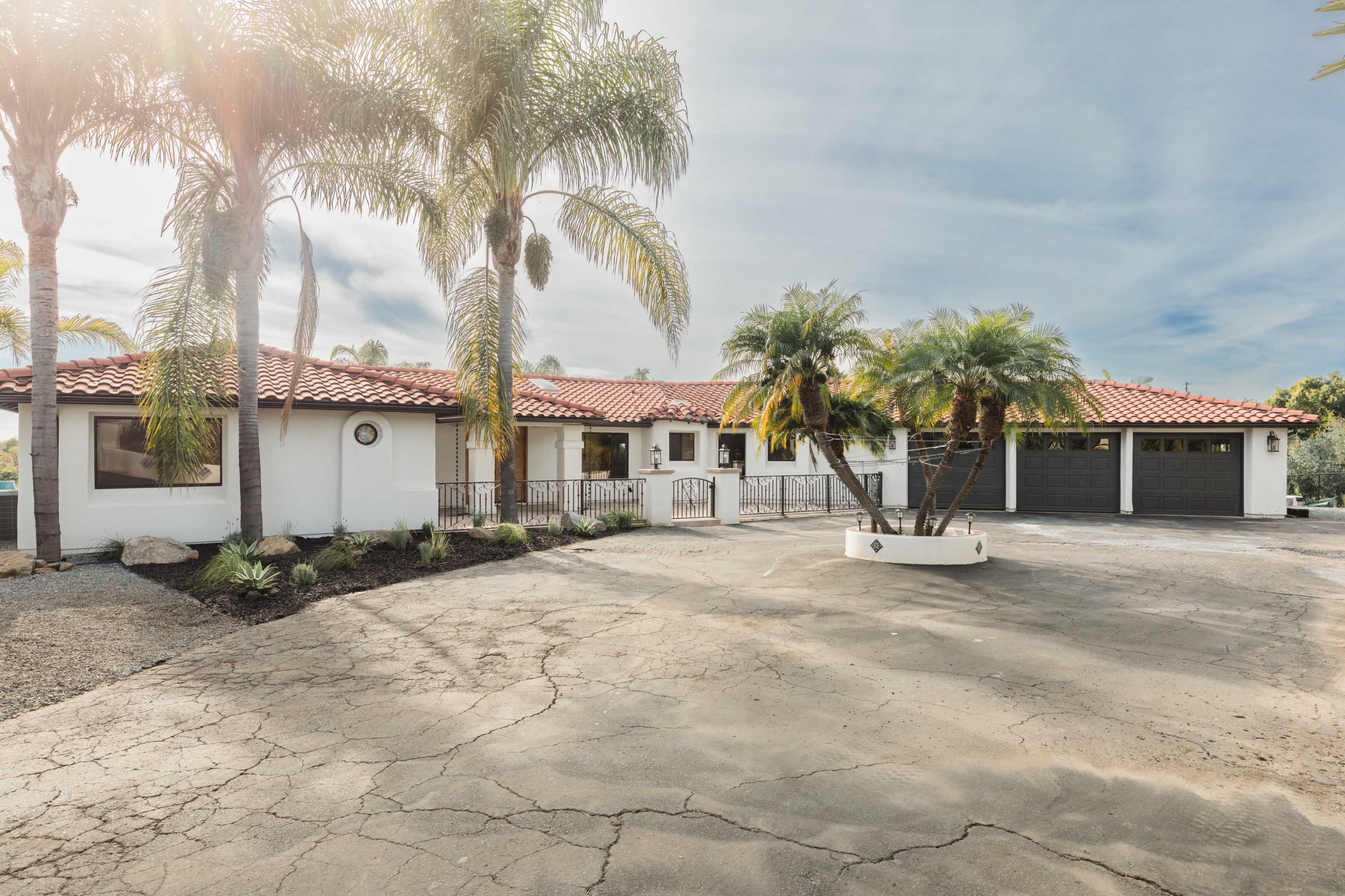 A single-story house with a red-tiled roof is surrounded by palm trees and features a large, cracked driveway.