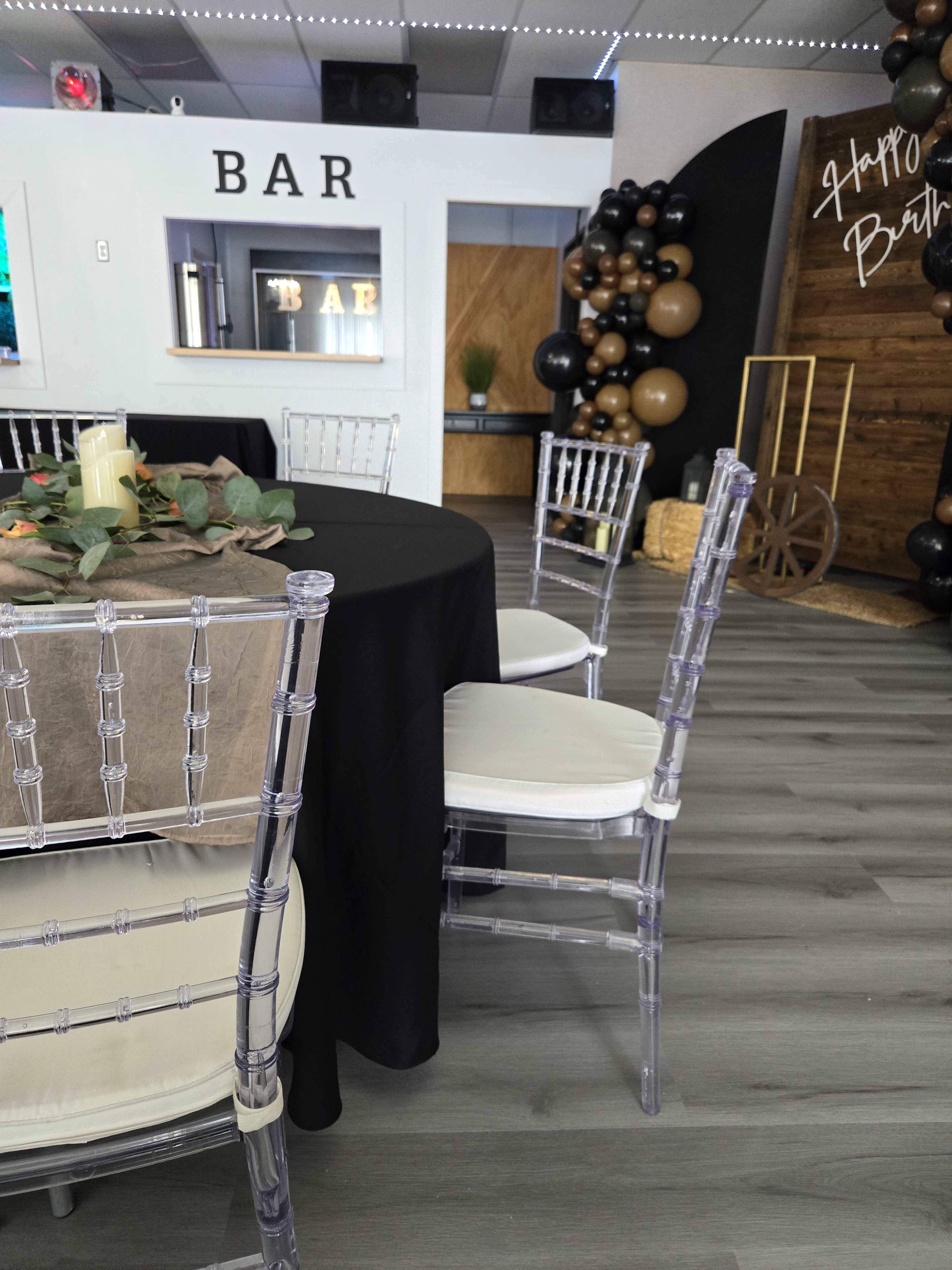 A decorated event space featuring a black table with a centerpiece, clear acrylic chairs, and a bar in the background, accented by black and gold balloons.