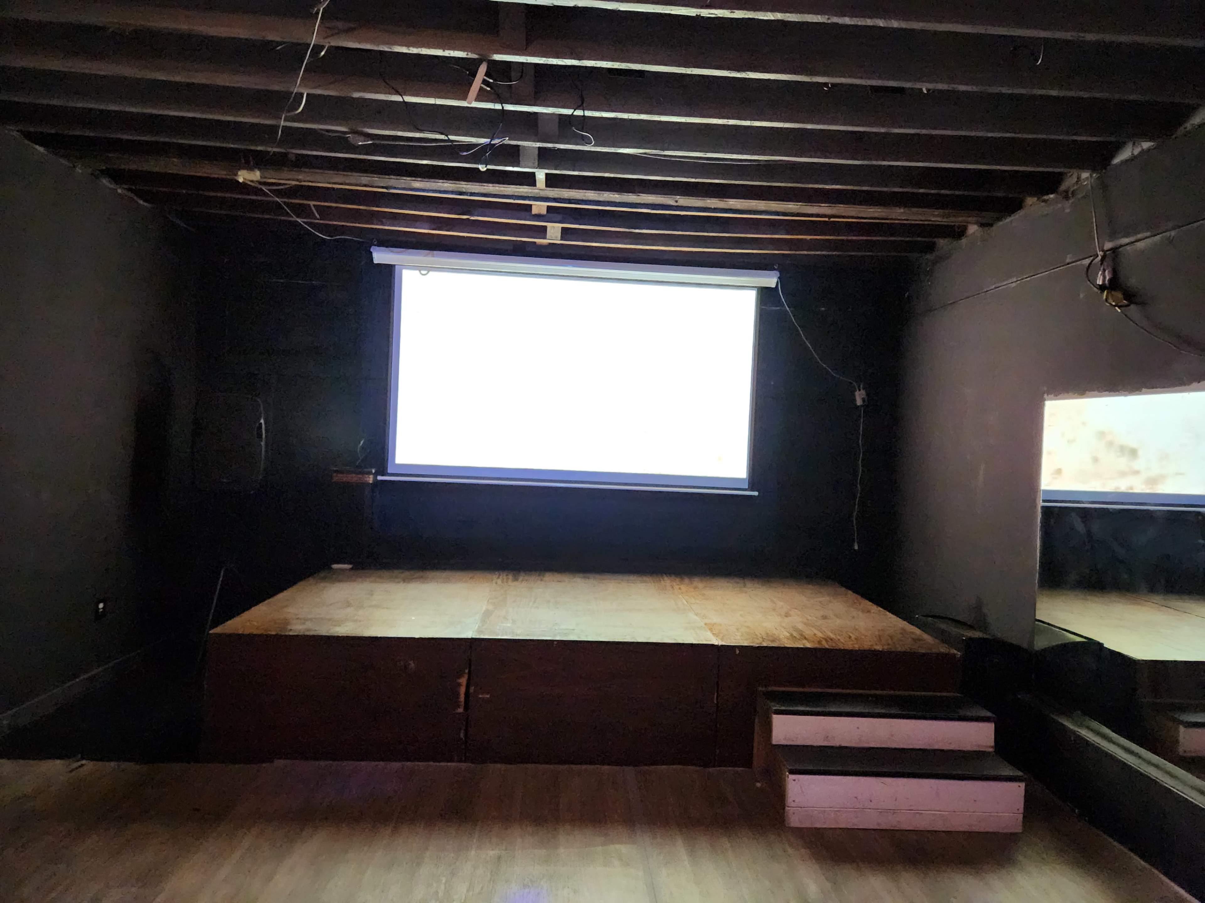 The image shows a dimly lit room with a wooden stage and a projector screen mounted on the wall.