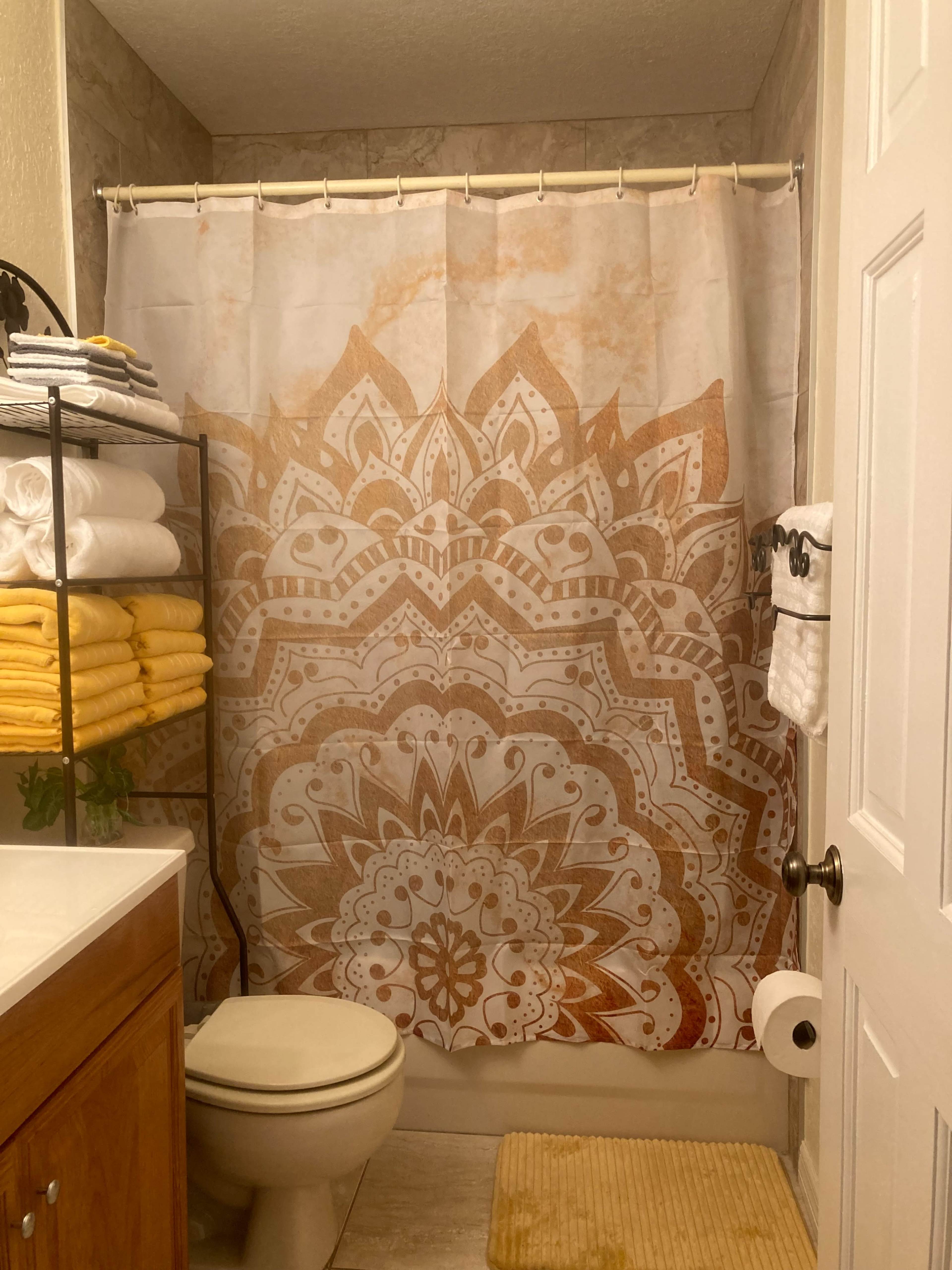 The bathroom features a shower with a mandala-patterned curtain, neatly stacked towels in various colors on a shelf, and a toilet beside a counter with a textured mat.