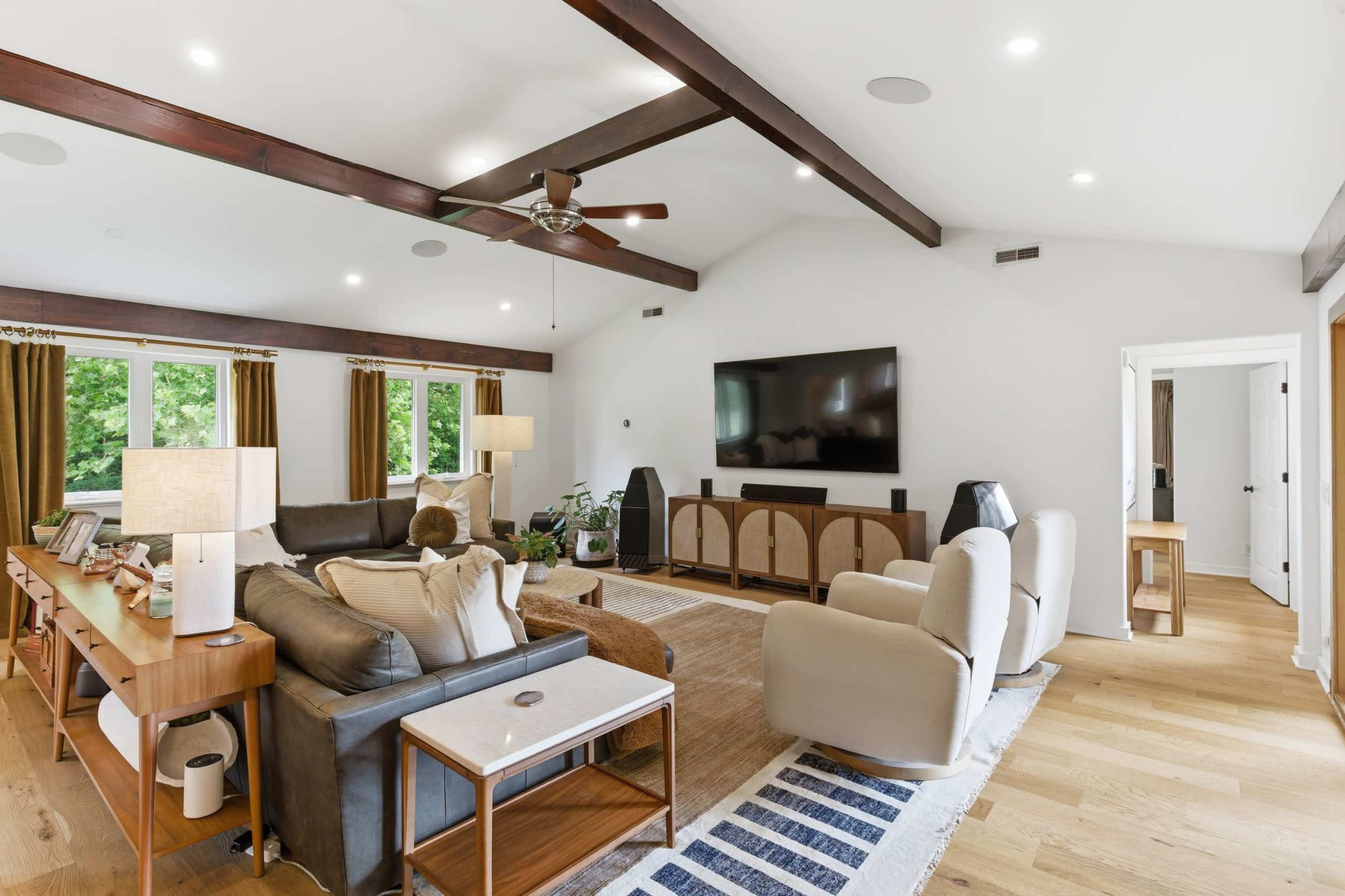The image shows a spacious living room featuring a large sectional sofa, a mounted flat-screen TV, and wood beams on the ceiling, with large windows allowing natural light to fill the space.