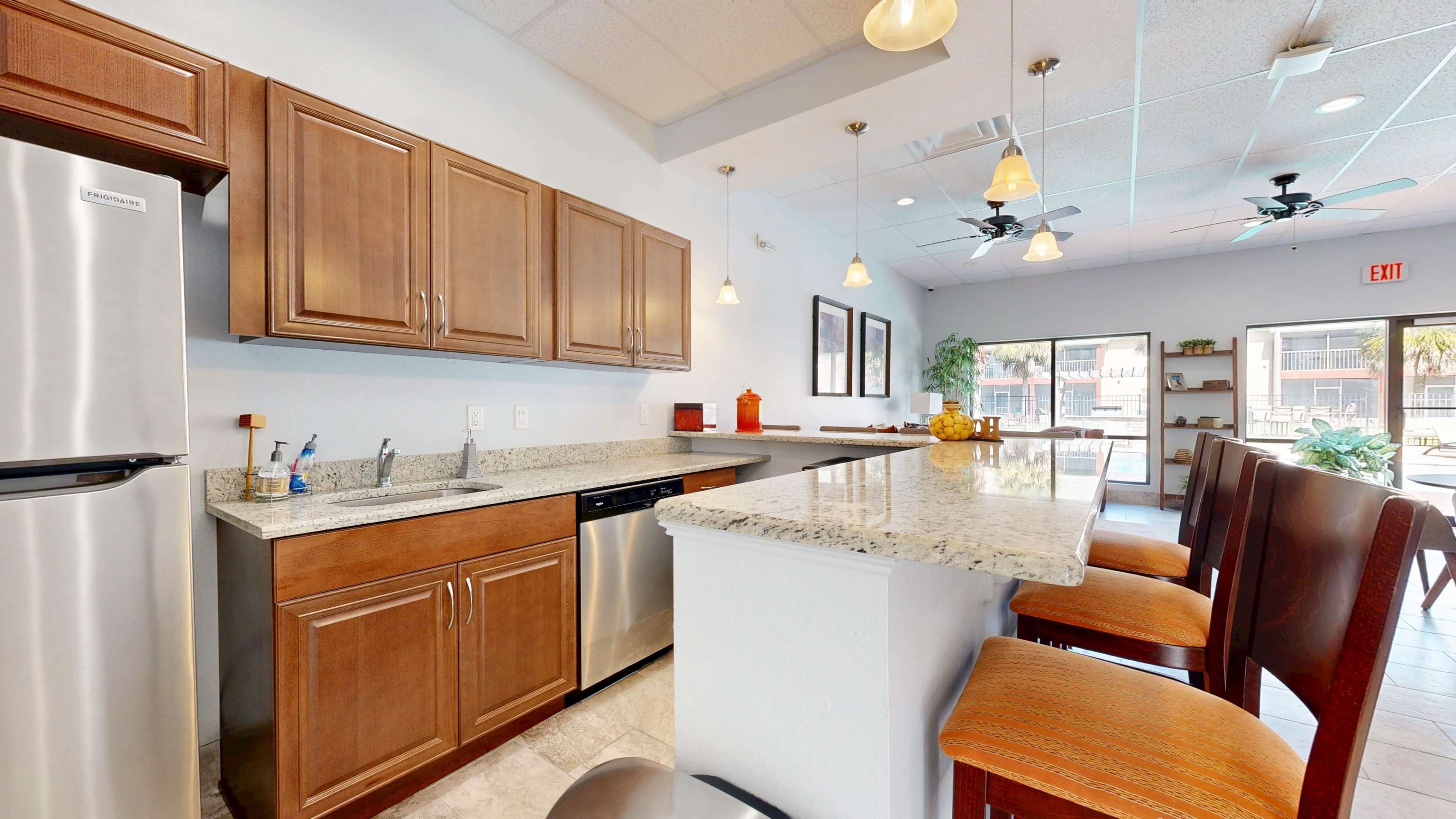 The image shows a modern kitchen area with wood cabinetry, stainless steel appliances, a granite countertop, and bar seating.
