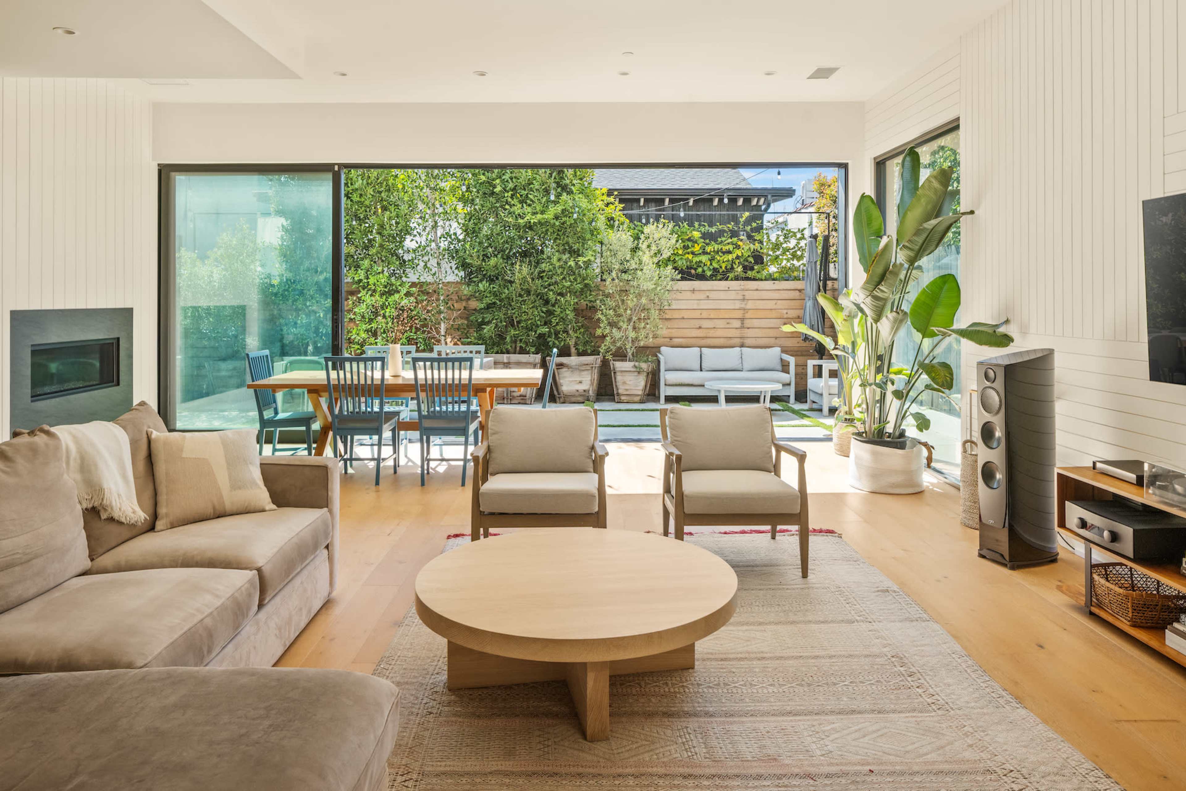 A modern living room featuring a beige sectional sofa, a round wooden coffee table, and large windows that open up to a backyard with a dining area and greenery.
