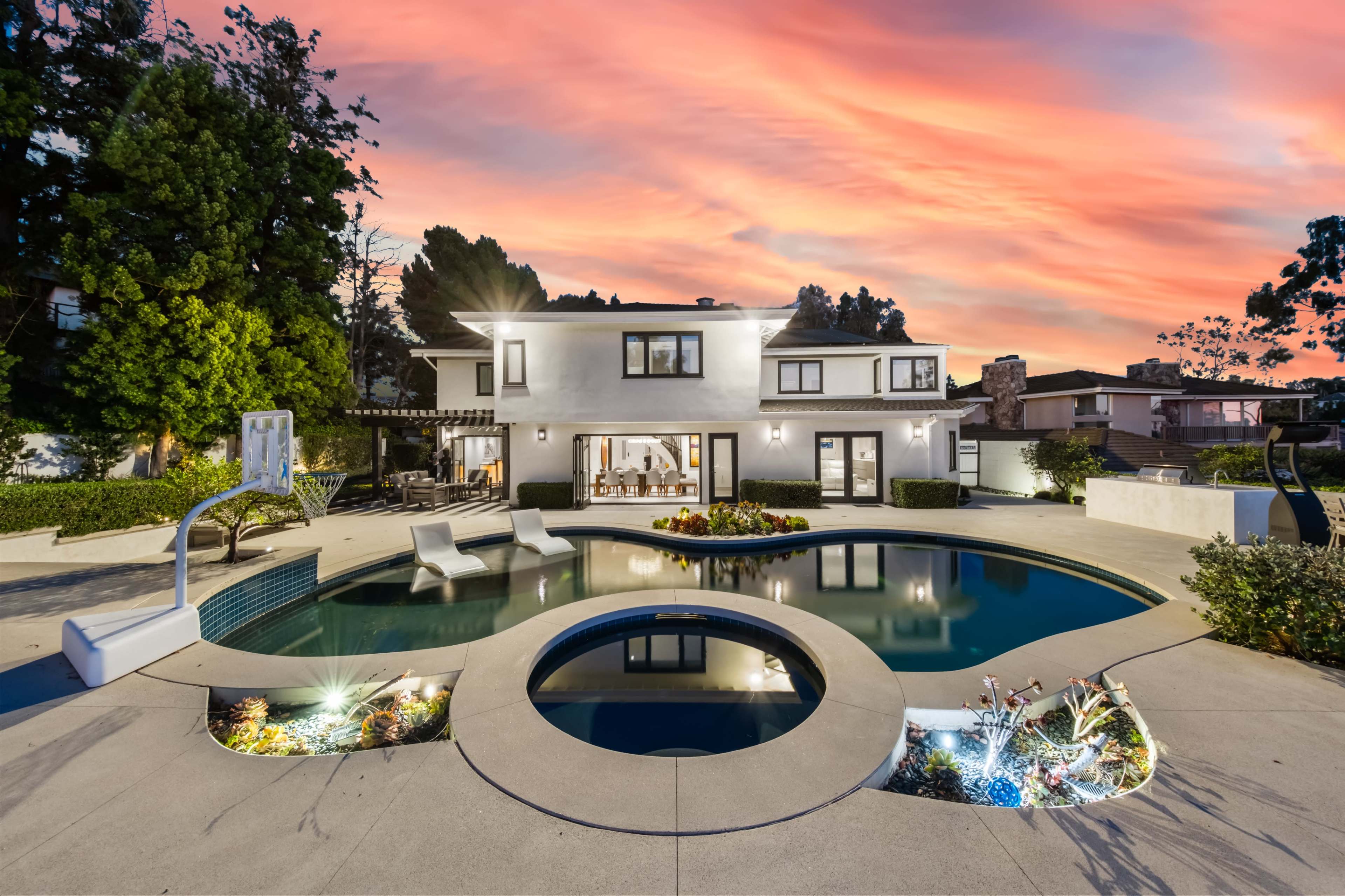 A modern two-story house with a pool features an outdoor seating area and well-maintained landscaping under a vibrant sunset sky.