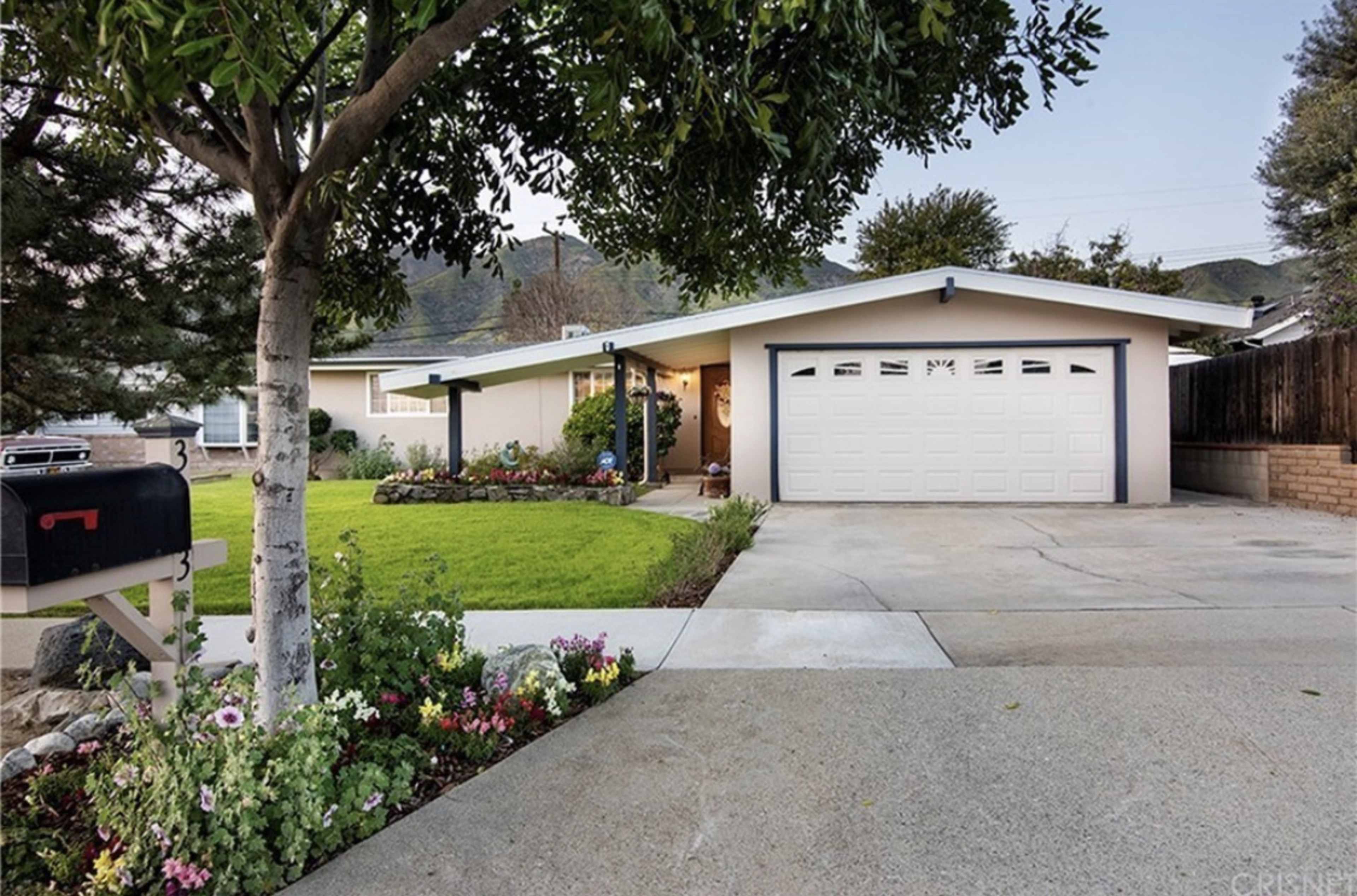 A single-story house with a two-car garage is set on a manicured lawn, surrounded by flower beds and a tree.