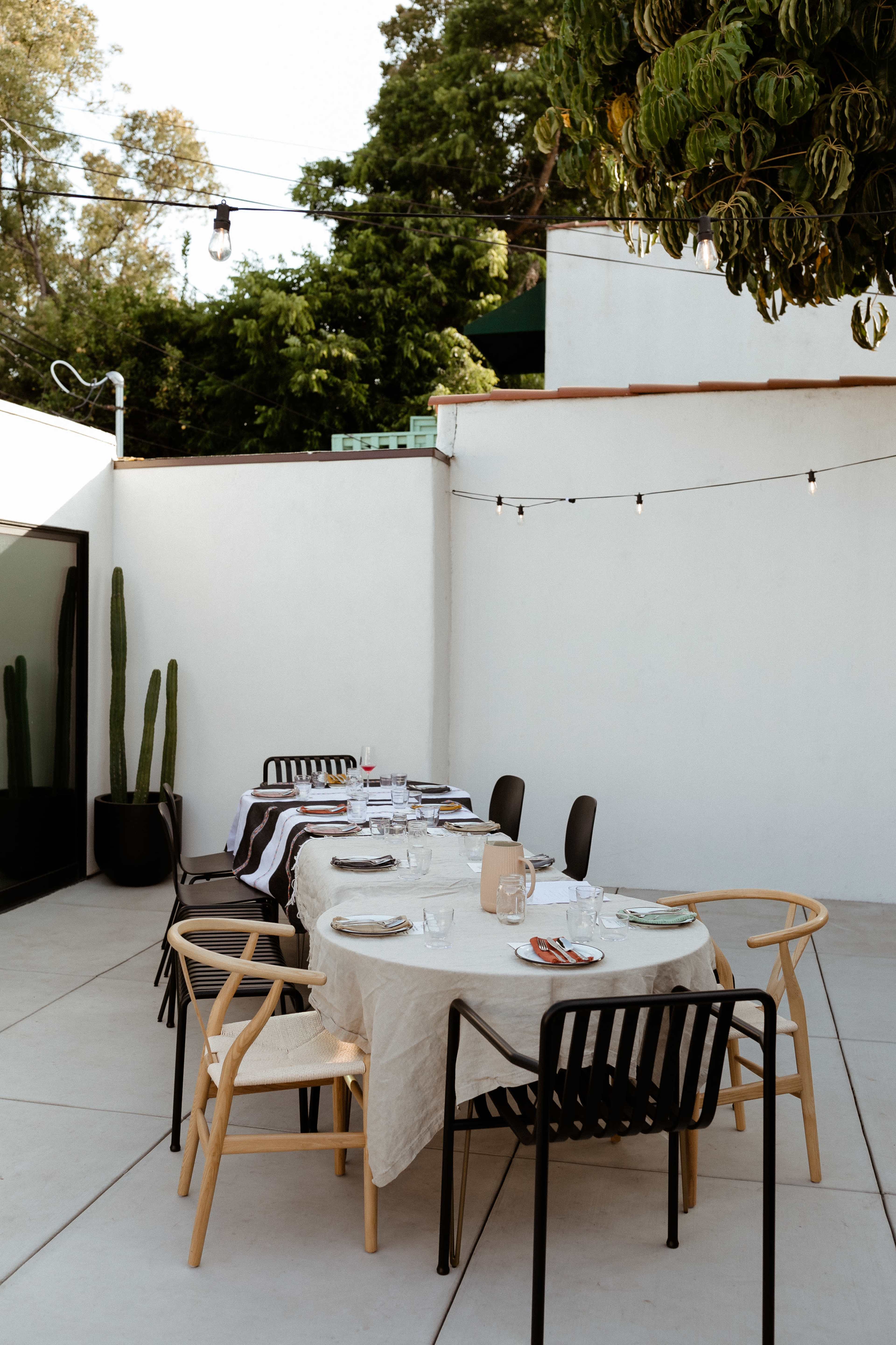 A long dining table is set up outdoors with dishes and glasses in a modern courtyard surrounded by greenery and string lights.