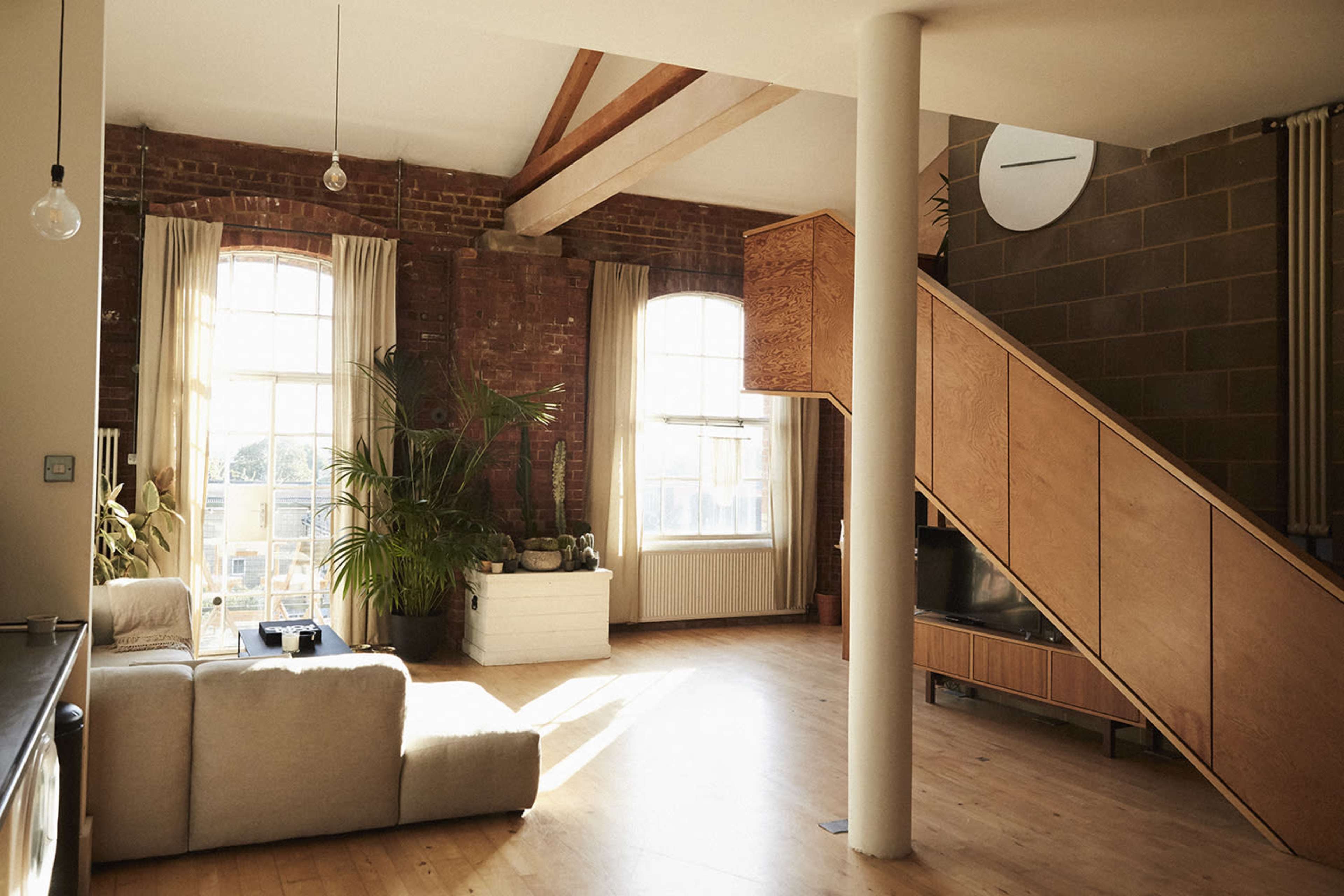 A spacious living area with large windows, exposed brick walls, and a wooden staircase.