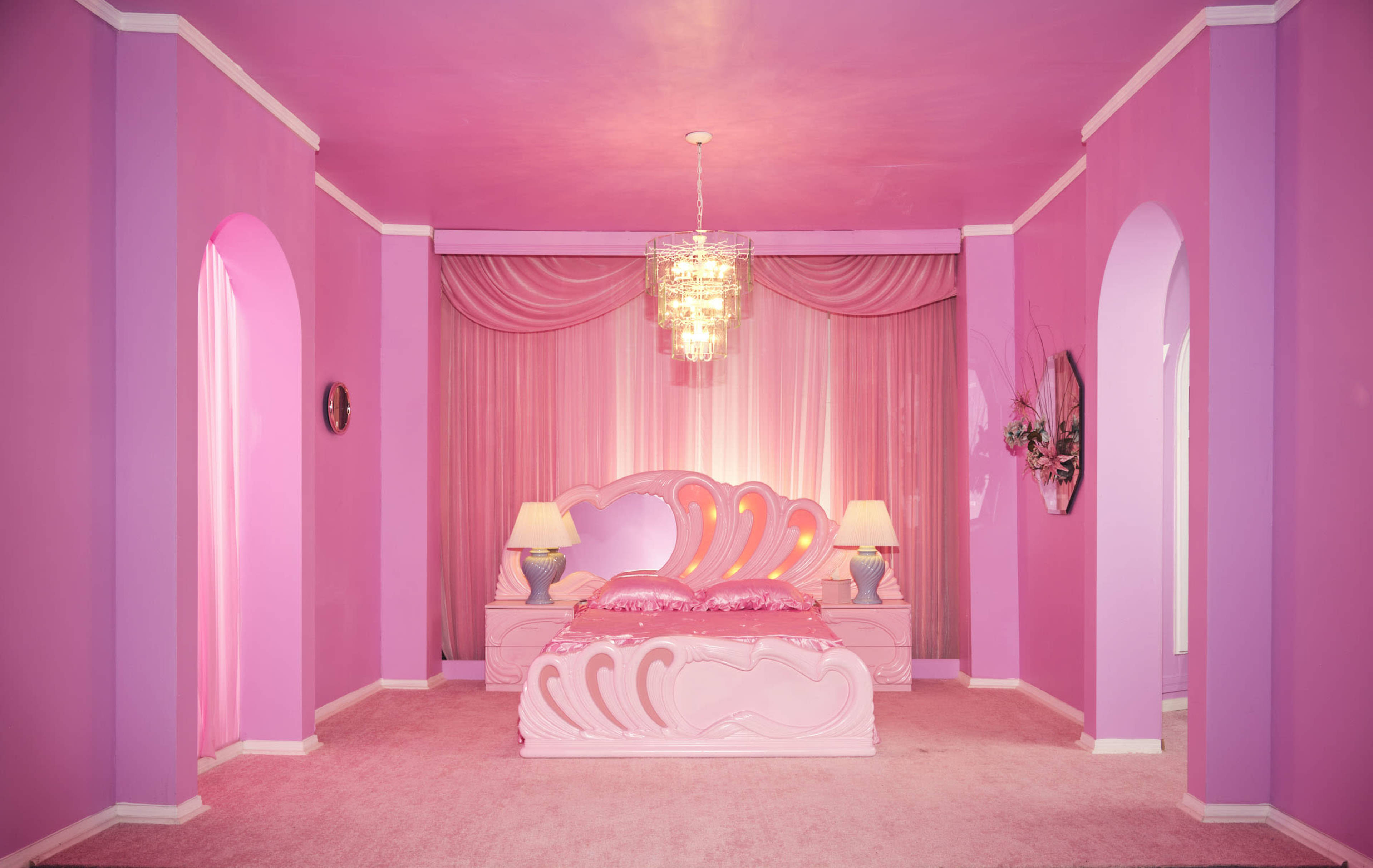 A bright pink bedroom features a decorative bed, matching lamps, and a chandelier, with draped fabric on the walls.