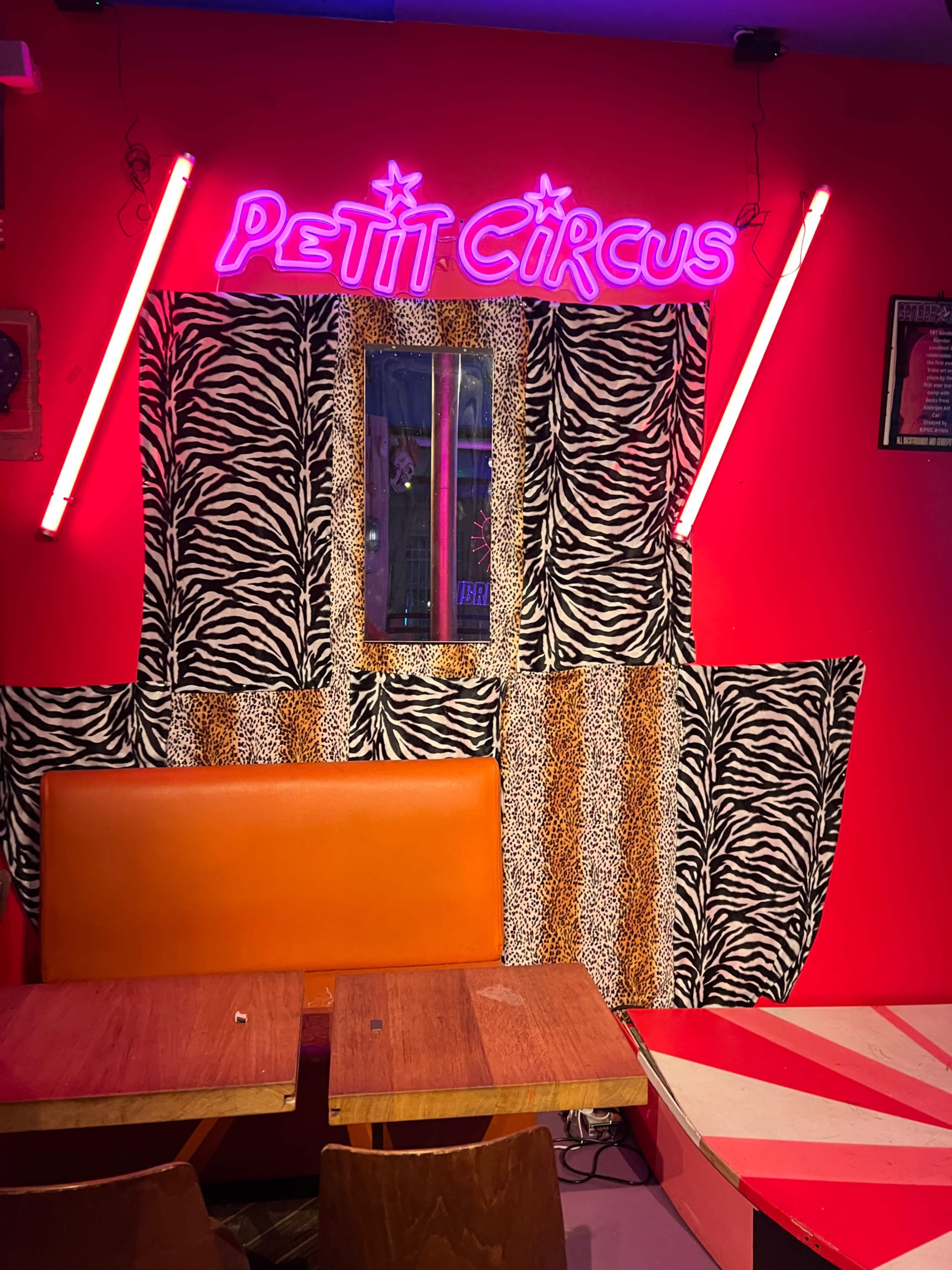 The image shows a brightly colored interior of a venue with a neon sign that reads "PETIT CIRCUS," zebra-patterned fabric, and orange seating.