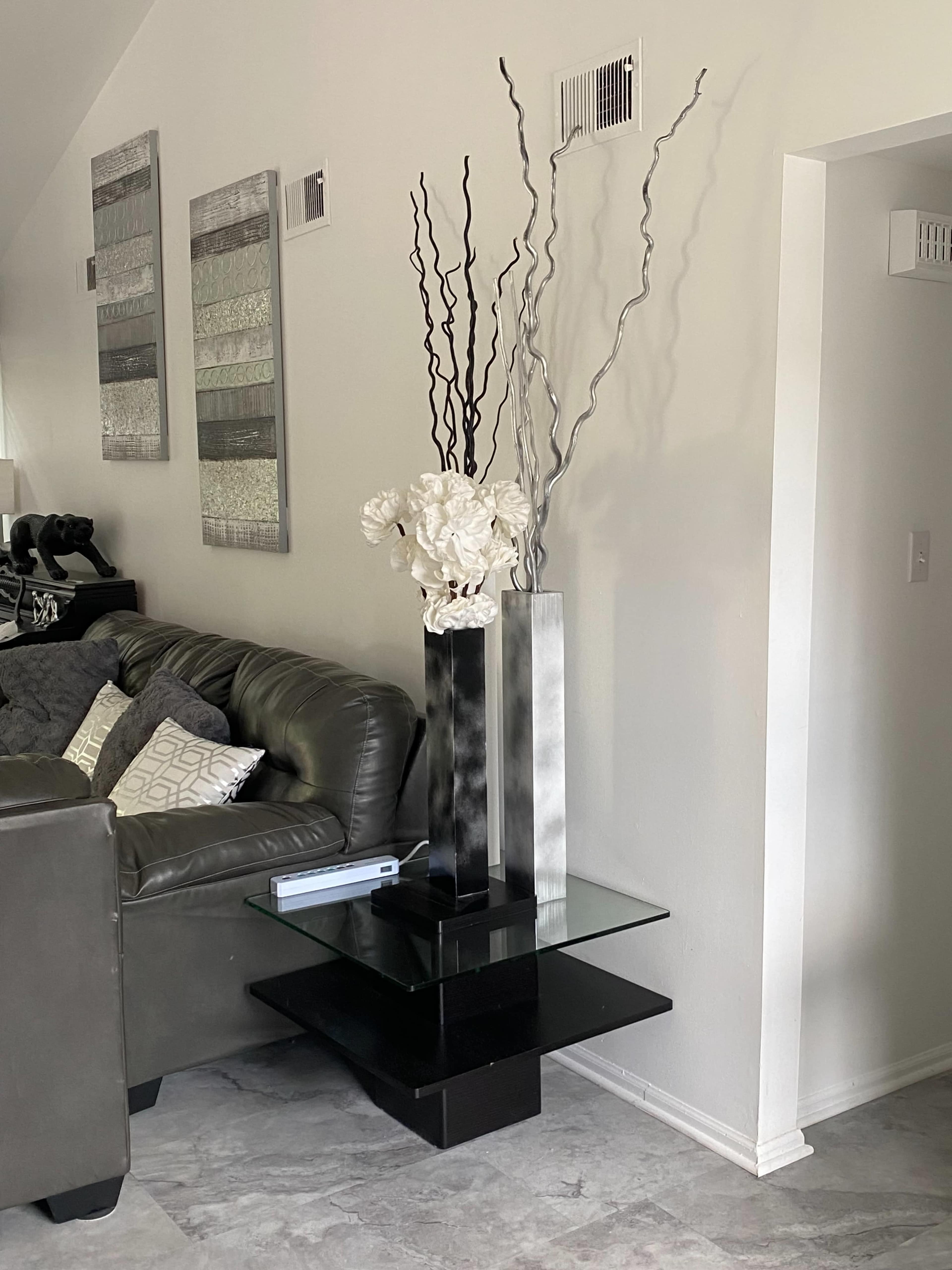 A modern living room corner features a black and glass accent table with a decorative sculpture and tall vases, accompanied by a gray sofa and textured wall art.