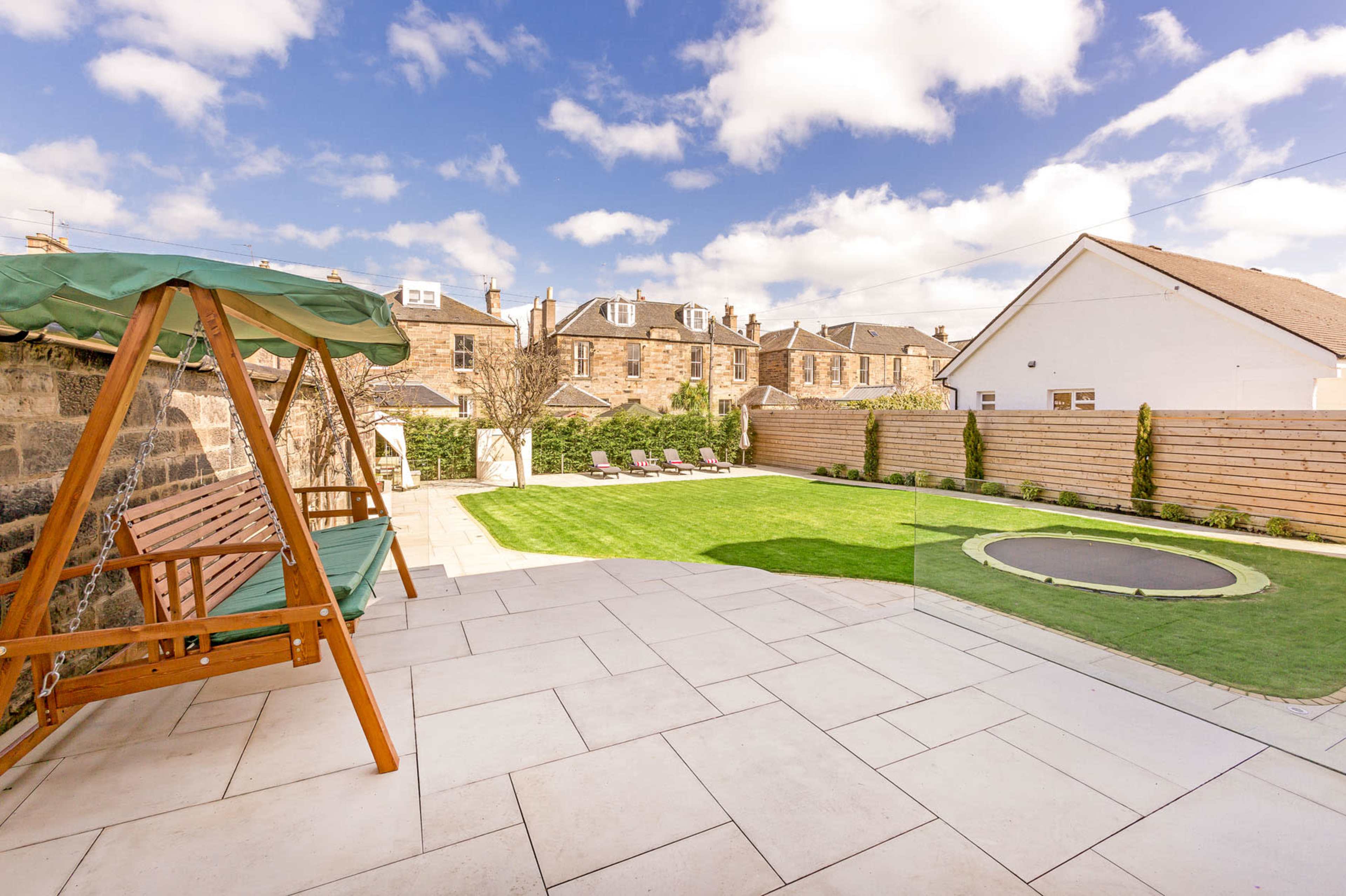 A backyard patio with a swing bench, neatly trimmed grass, a trampoline, and lounge chairs under a partly cloudy sky.