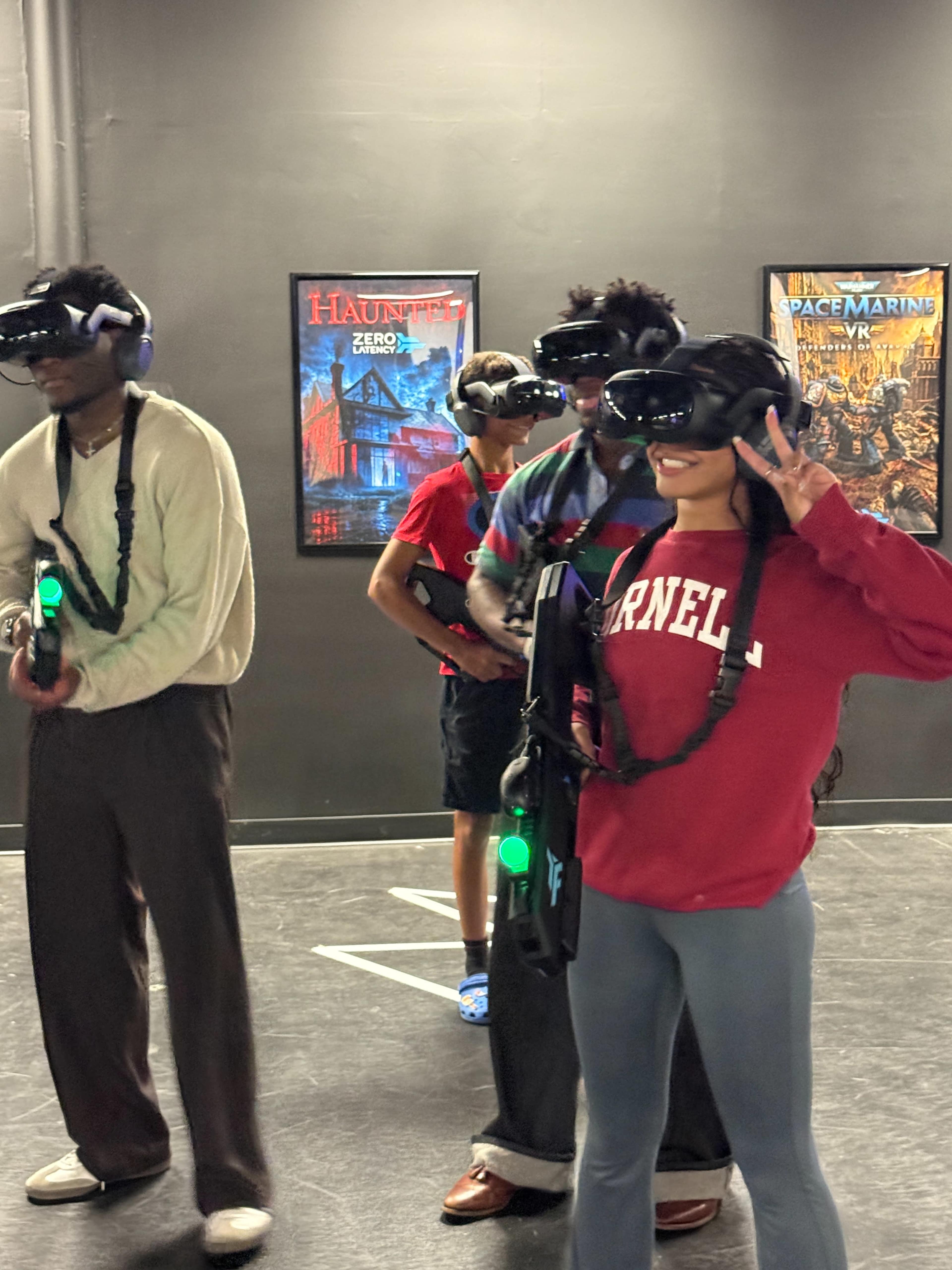 A group of four individuals wearing virtual reality headsets and holding gaming guns stands in a gaming space adorned with themed posters on the walls.