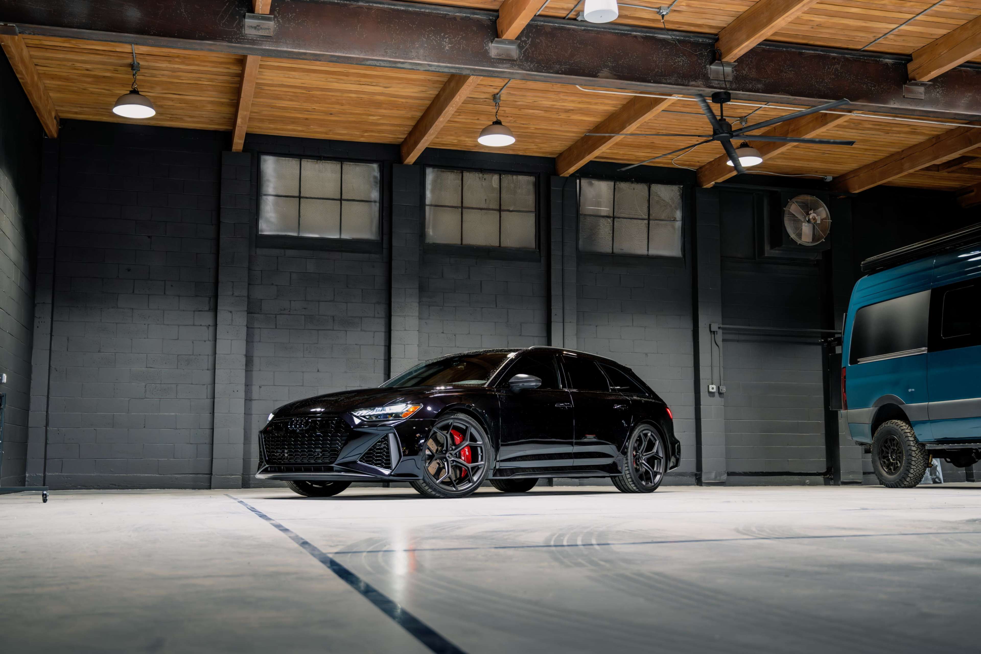 A black sport sedan is parked in a spacious garage with wooden beams and a blue van in the background.