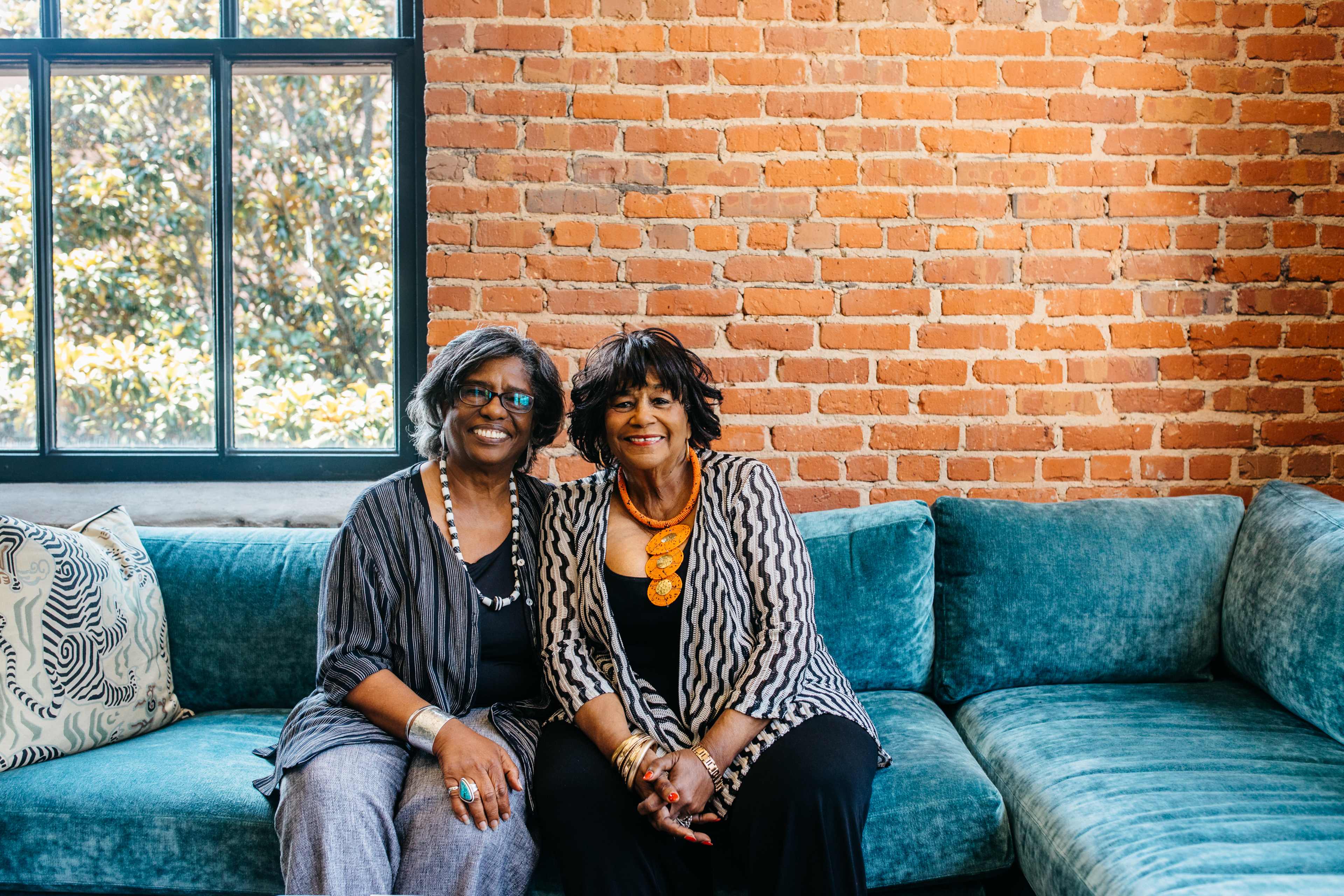 Two women sit on a teal sofa in front of a large window with brick walls in the background.