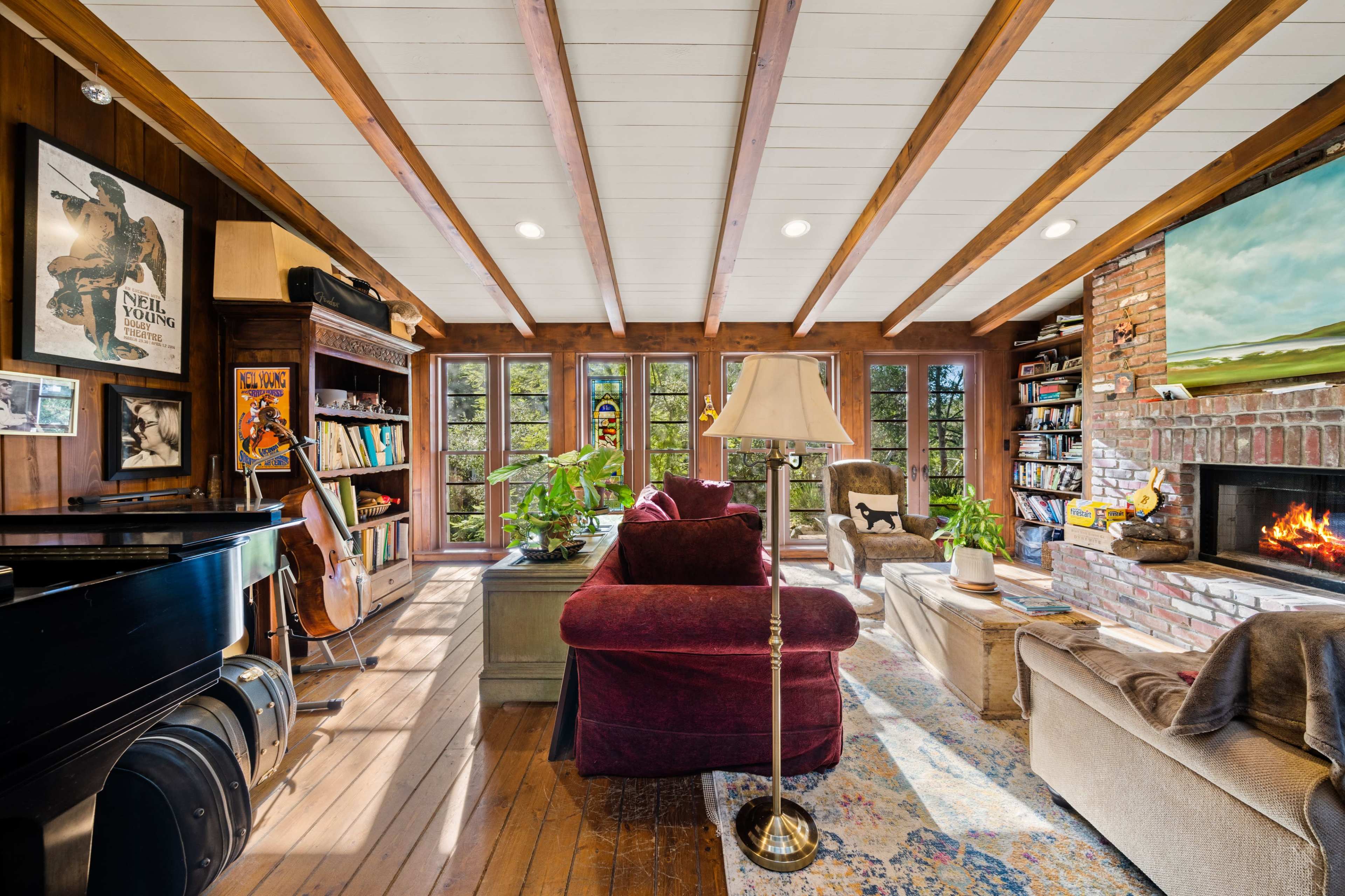 A cozy living room with wooden beams, a piano, bookshelves, a fireplace, and large windows that provide a view of the outdoors.