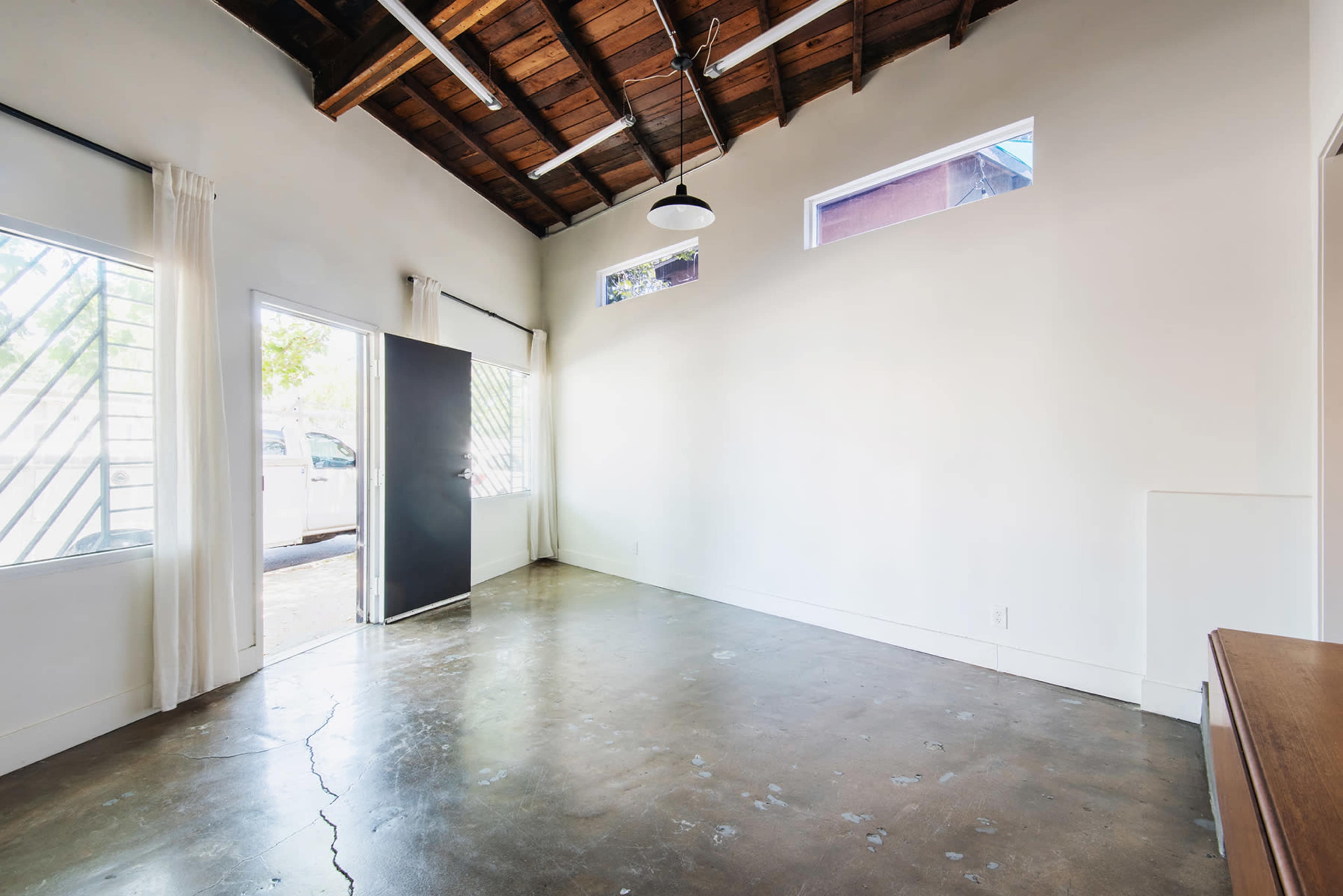 The image shows a minimalist room with a concrete floor, wooden ceiling beams, and a large open doorway leading outside.