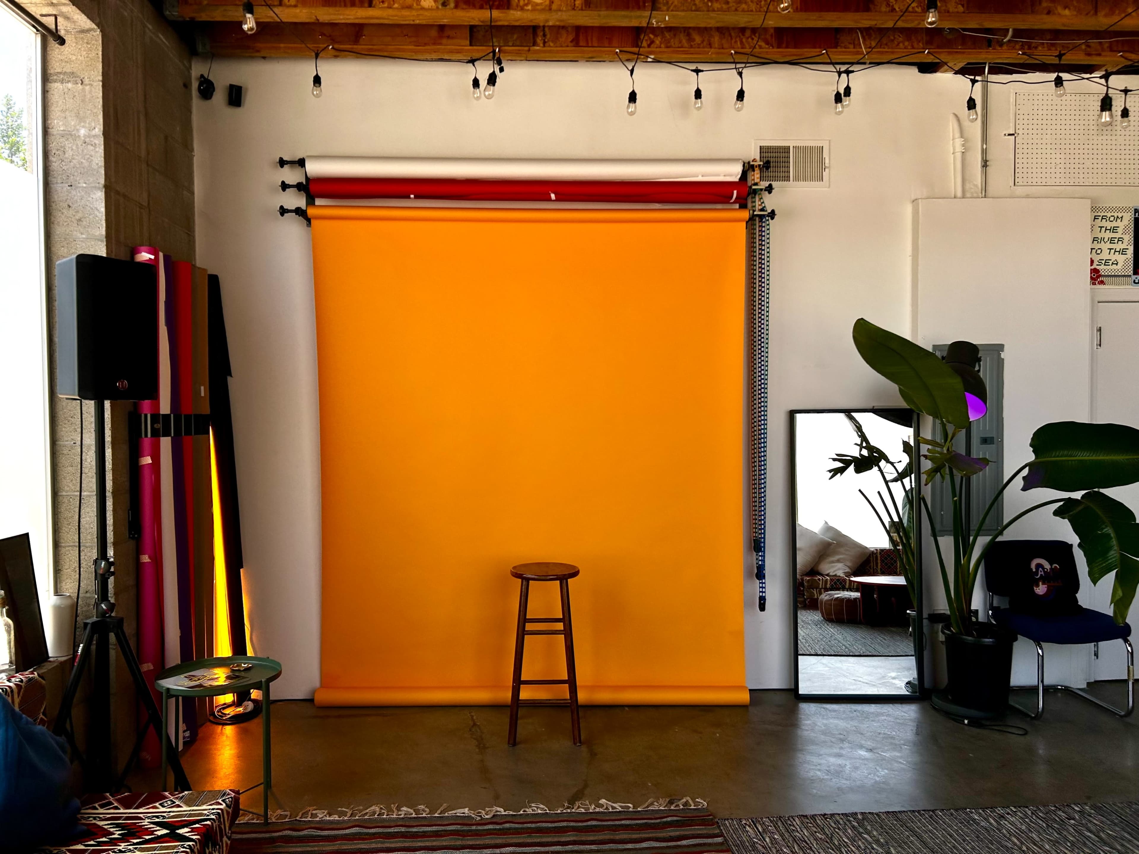 A studio space featuring a vibrant orange backdrop with a wooden stool in front of it, accompanied by a mirror and indoor plants.