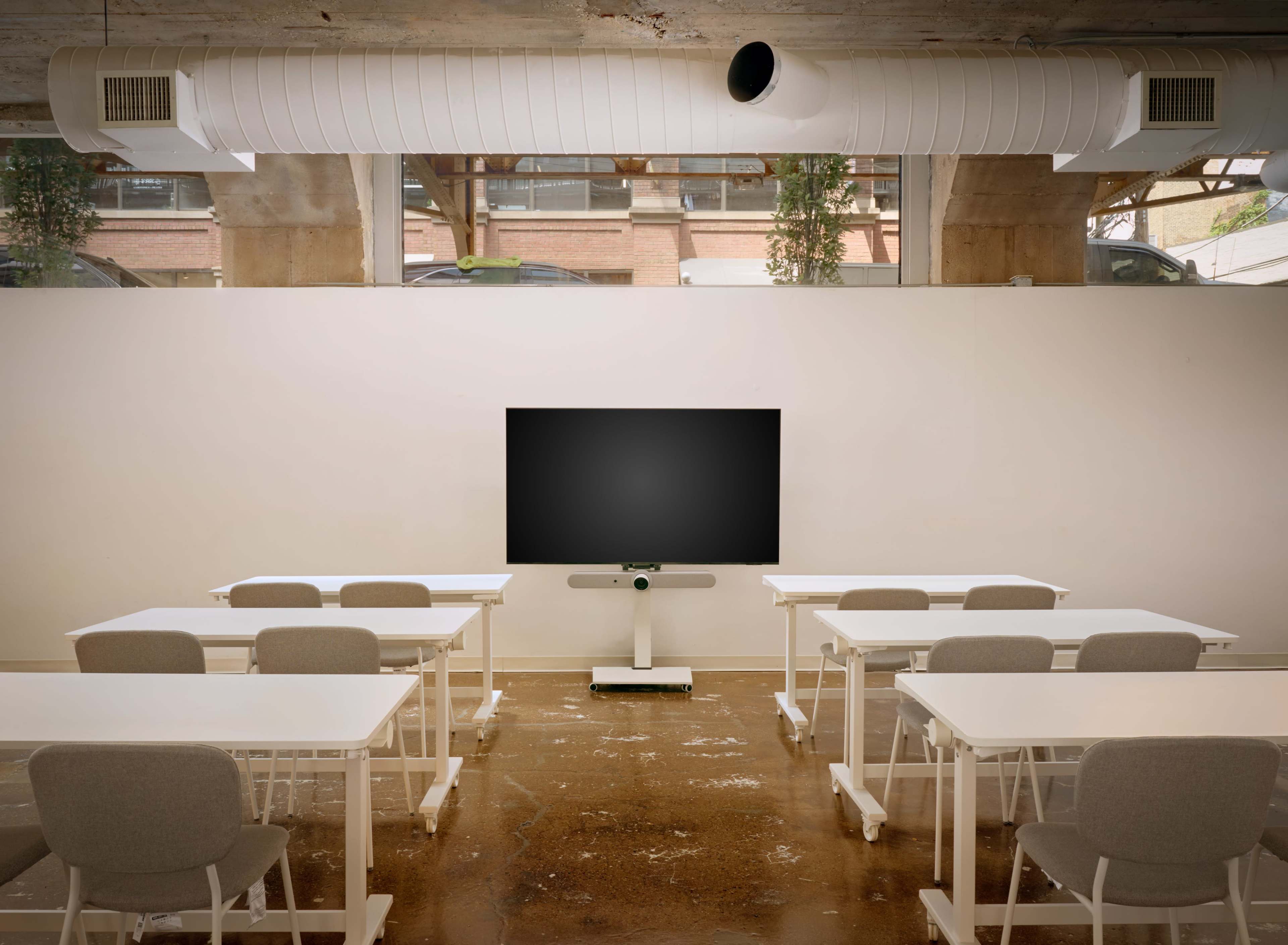 The image shows a classroom setting with several white desks and chairs facing a large blank wall-mounted screen.