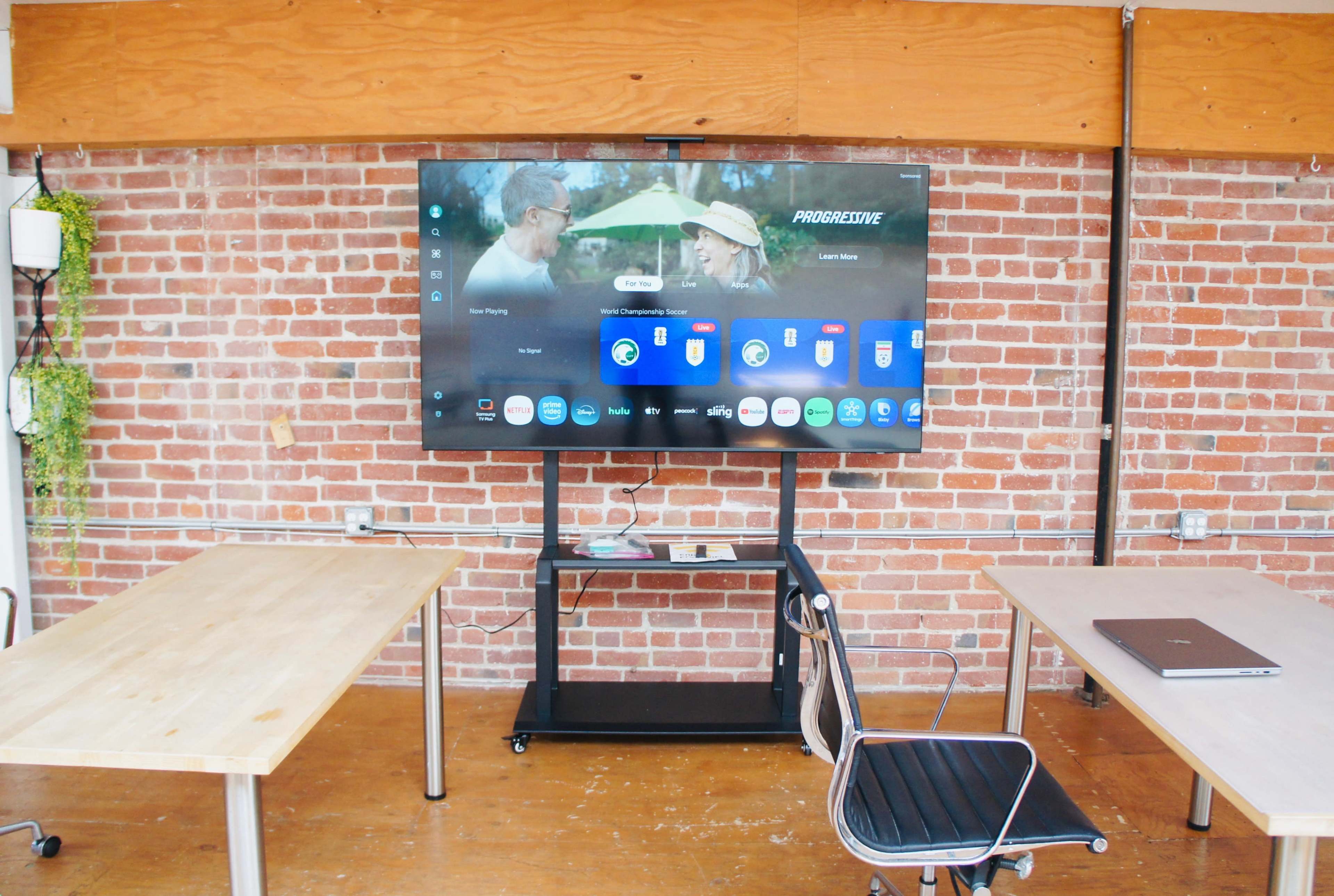 A large flat-screen TV is mounted on a brick wall, displaying a selection of app icons, while two wooden tables and a black chair are arranged nearby.