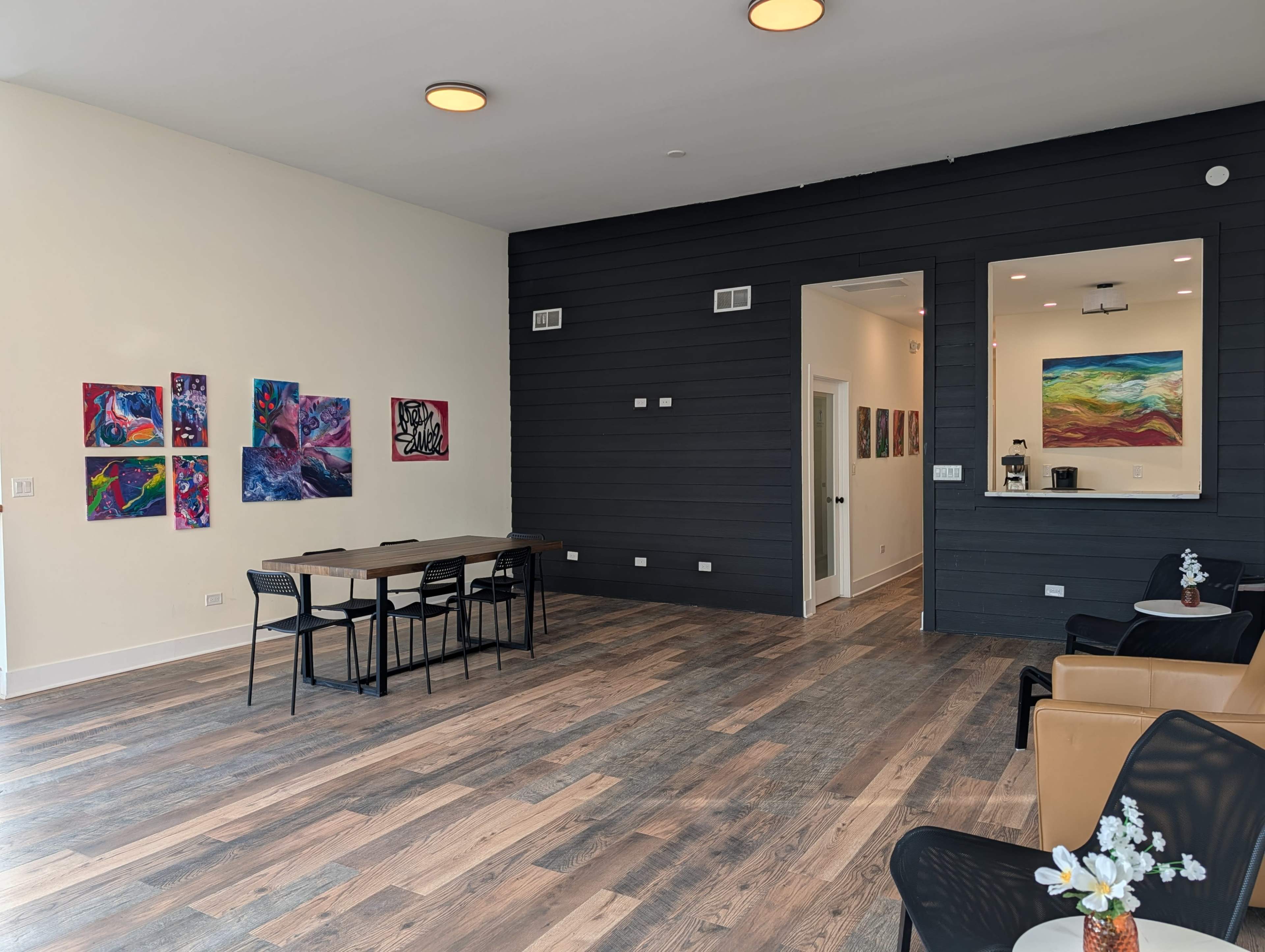 The image shows a spacious interior with a wooden floor, featuring a table with chairs, two seating areas, and walls adorned with colorful artwork.
