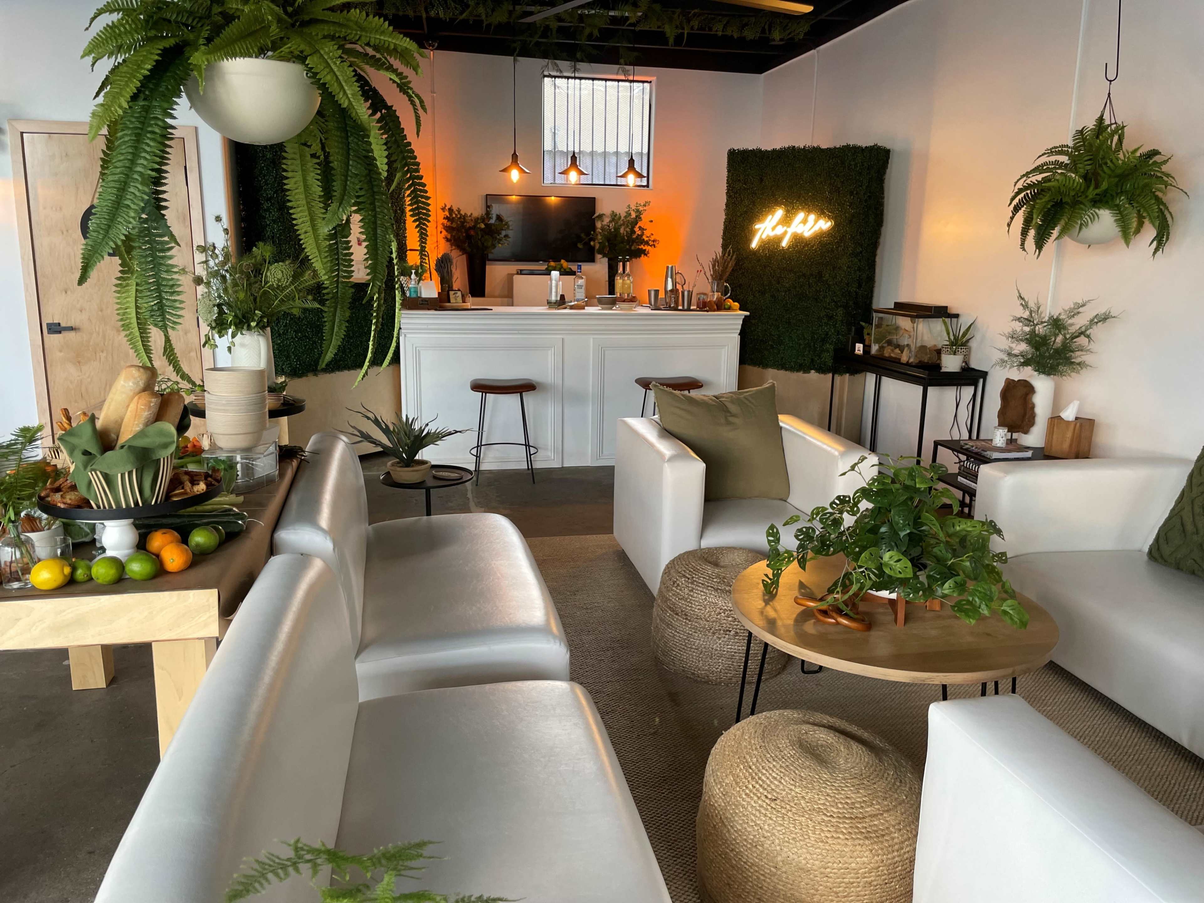 The image shows a modern, minimalist interior space featuring light-colored furniture, a bar area with plants, and decorative greenery on the walls.