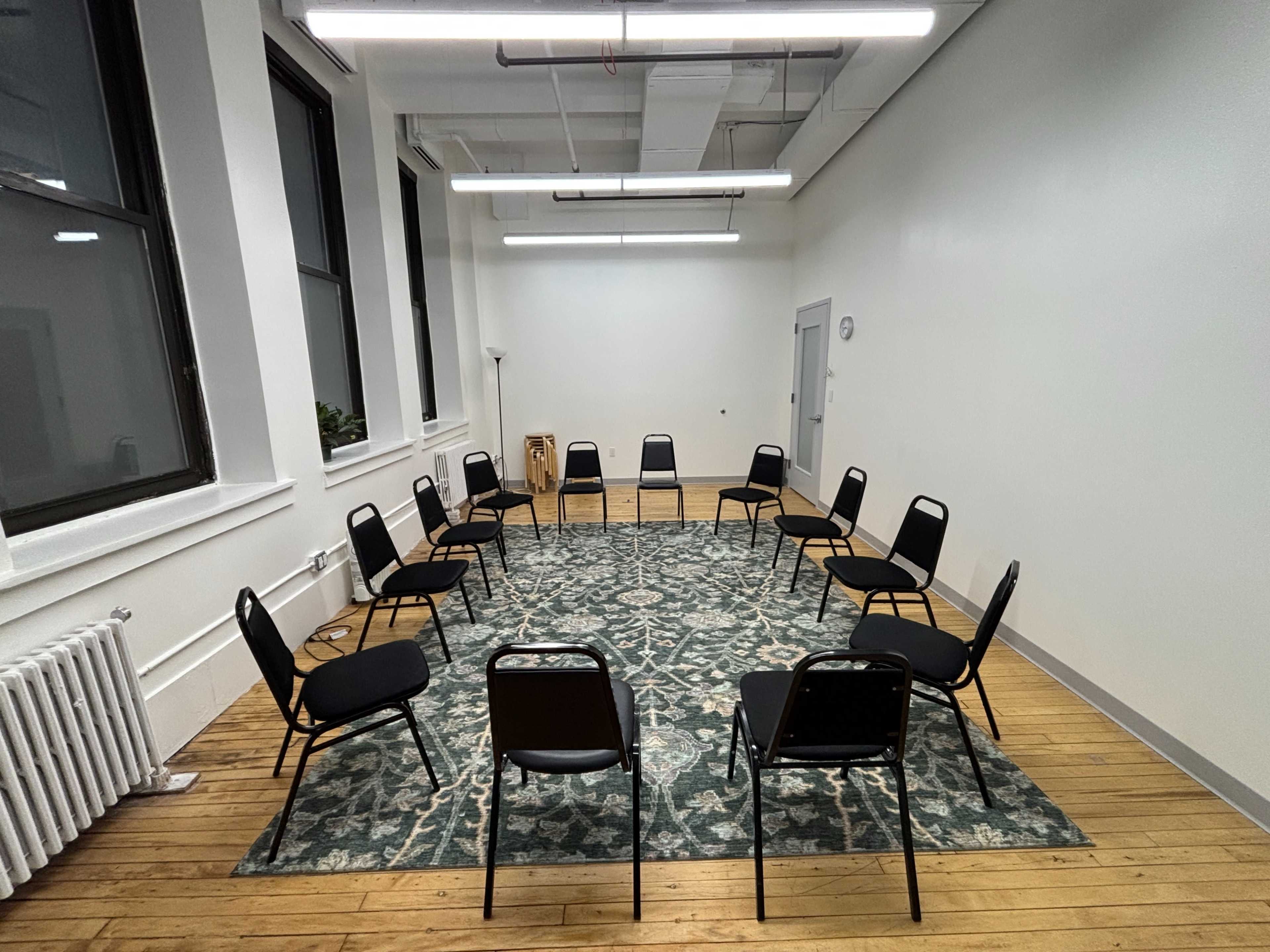 The image shows a spacious room with a circular arrangement of black chairs on a patterned rug.