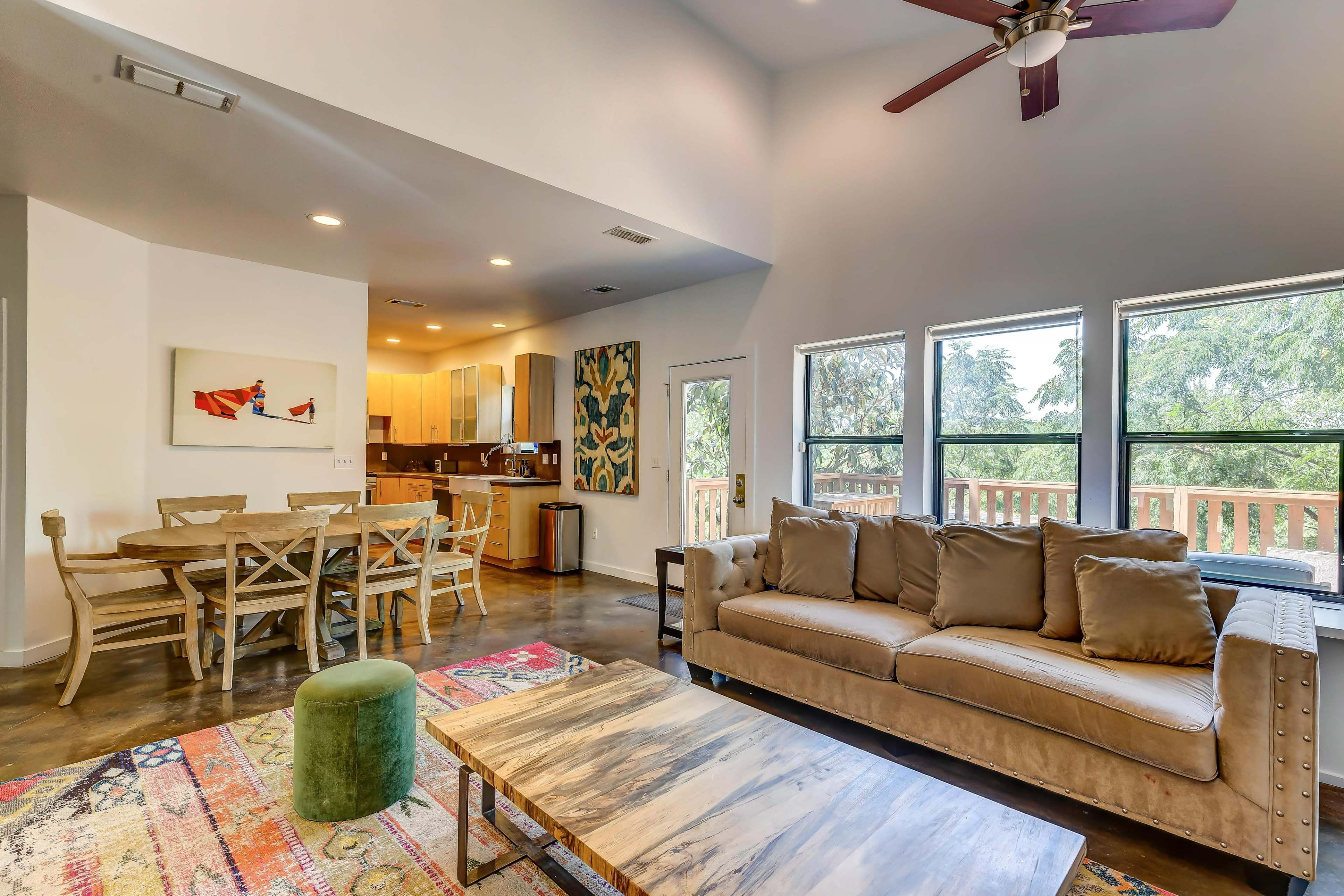 A modern living area featuring a beige sofa, a wooden dining table, and large windows that provide natural light, overlooking a deck.